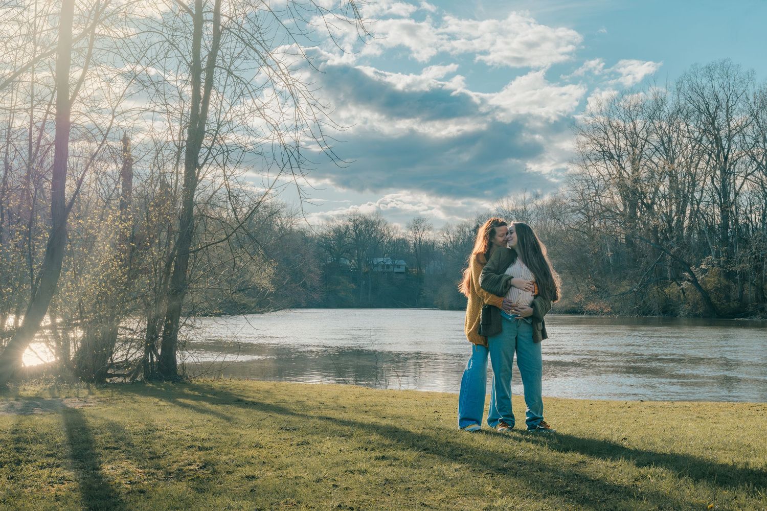 A couple embraces by a lake at sunset with bare winter trees in the background.