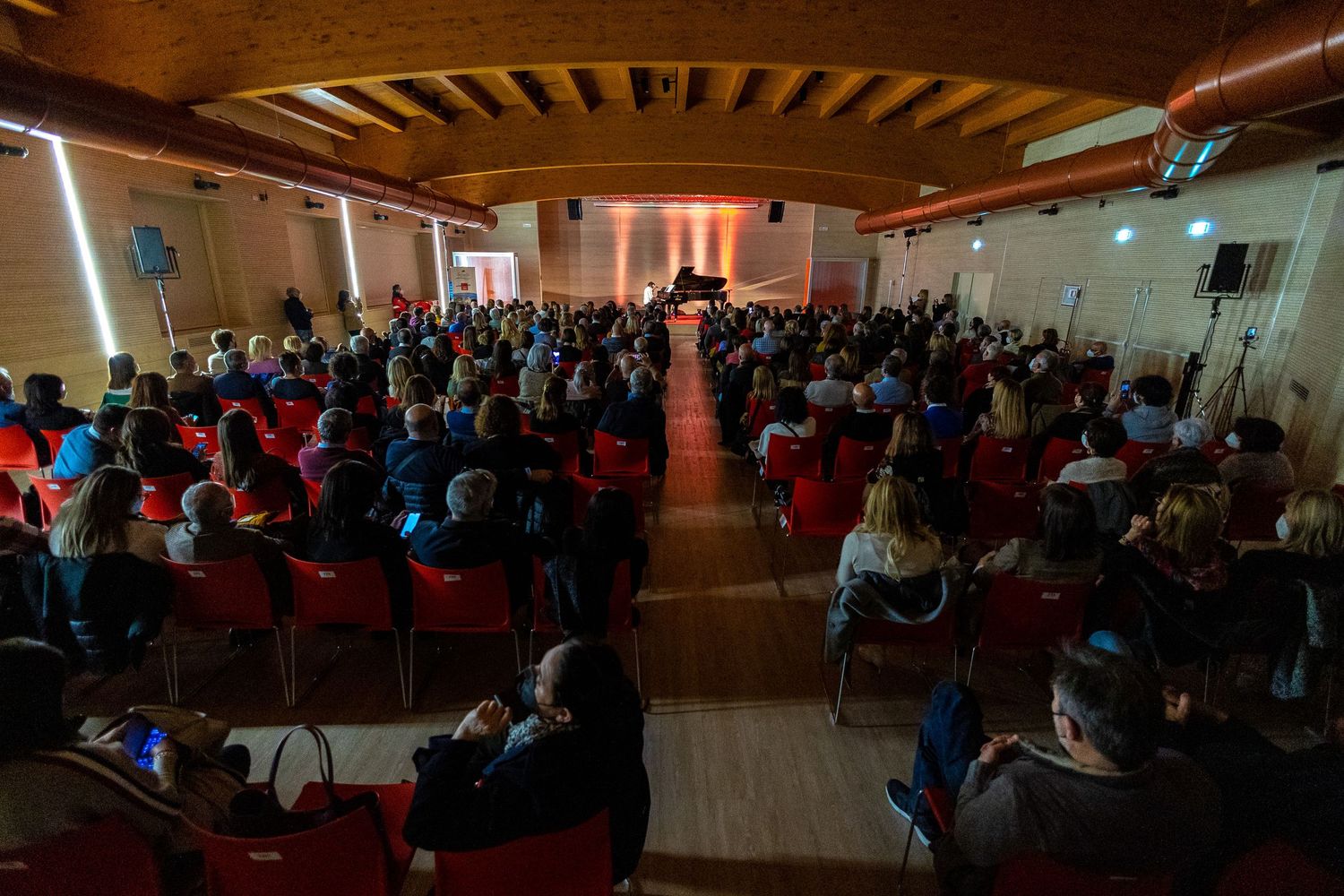 Crowded indoor auditorium filled with seated attendees under wooden beam ceiling.