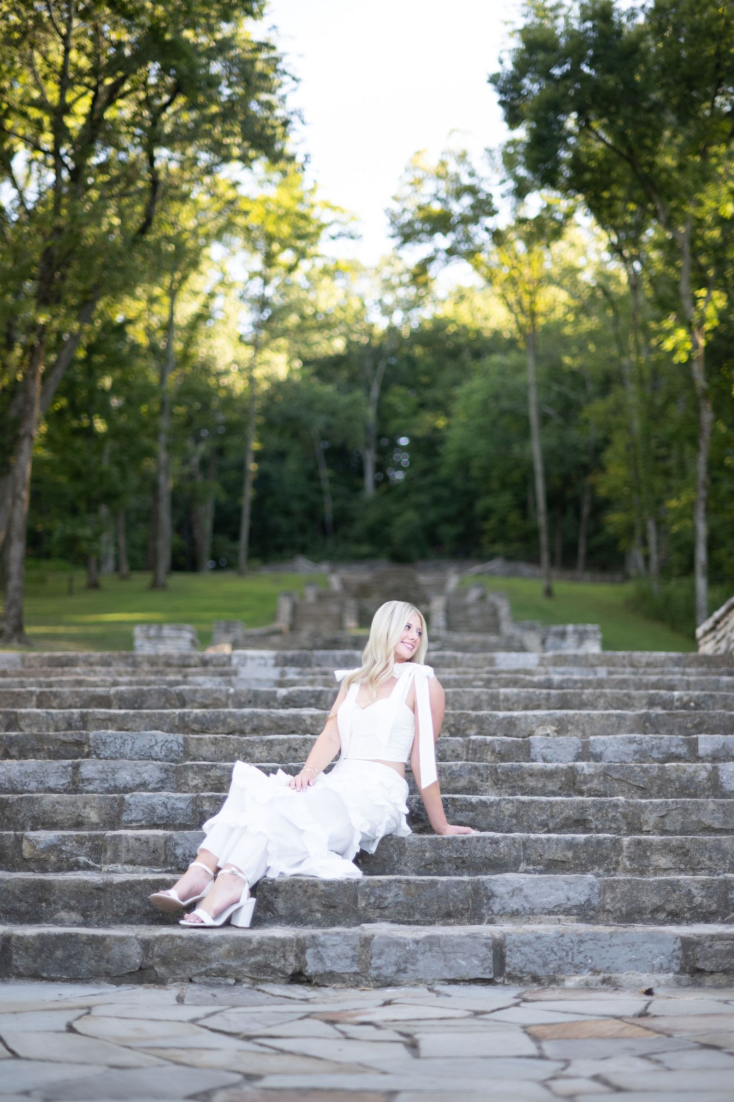 A figure in a white dress reclines on stone steps in a wooded outdoor amphitheater setting.