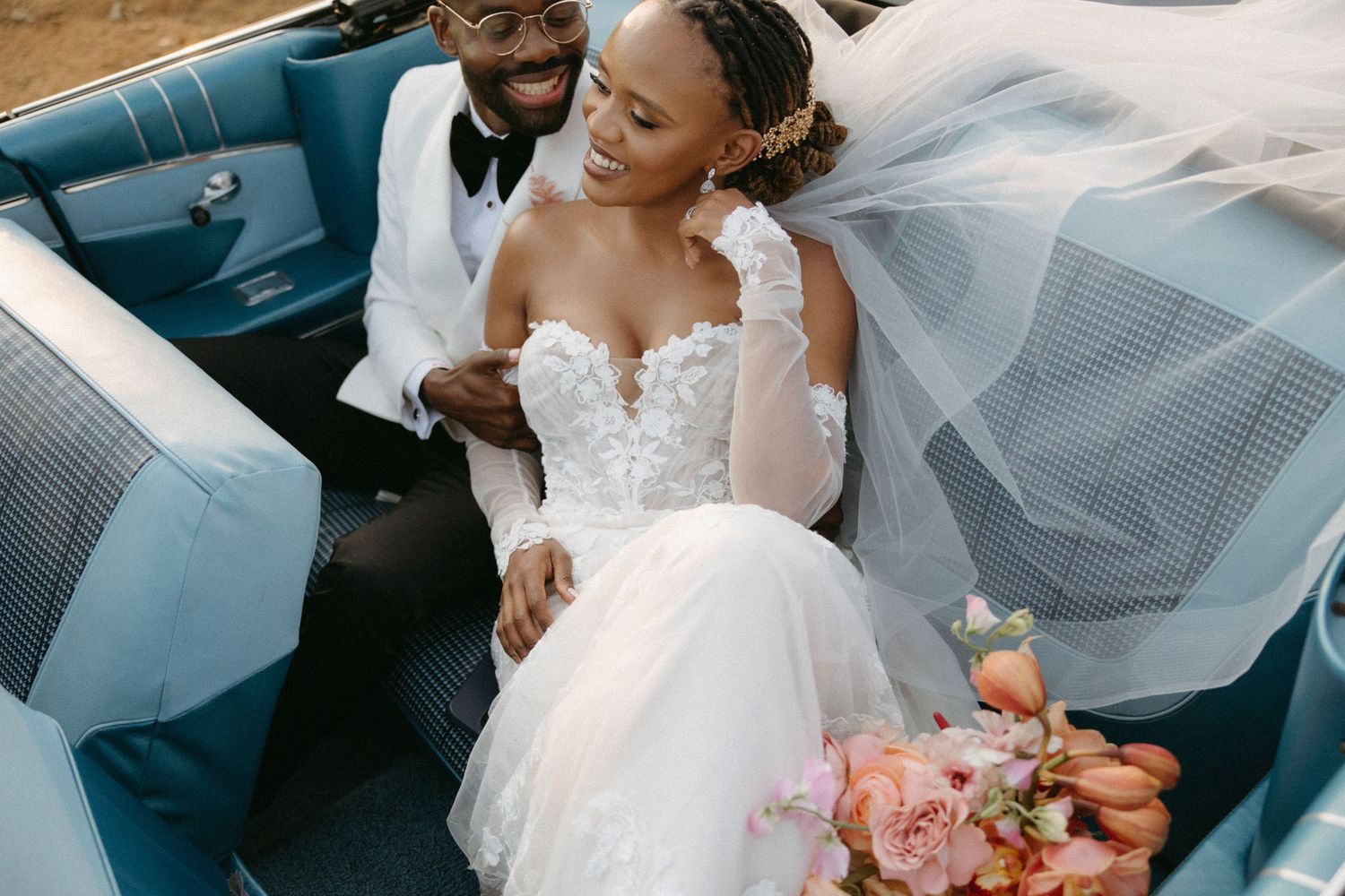 Bride and groom laughing in a vintage blue convertible, bride in lace gown with veil, holding a pink bouquet.