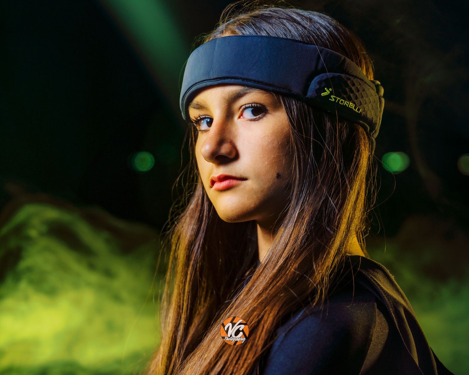 Young woman wearing a navy blue Storelli headband and sports jersey, posed against a dramatic green smoky background.