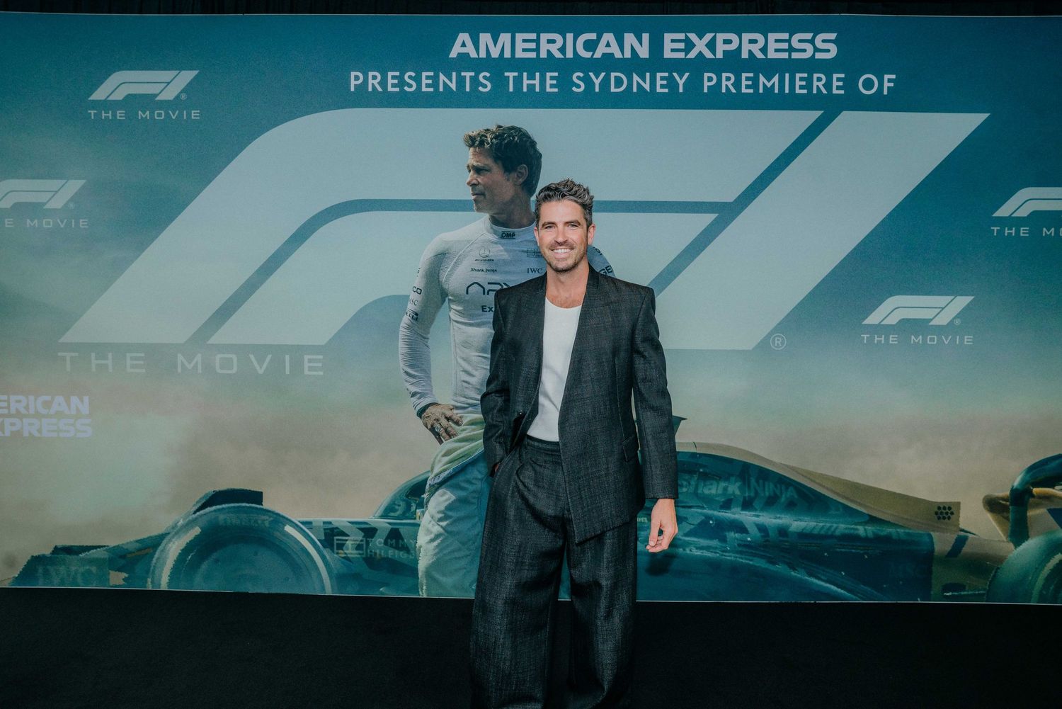 A man in a black suit poses at the American Express Sydney Premiere of F1 The Movie in front of a promotional backdrop.