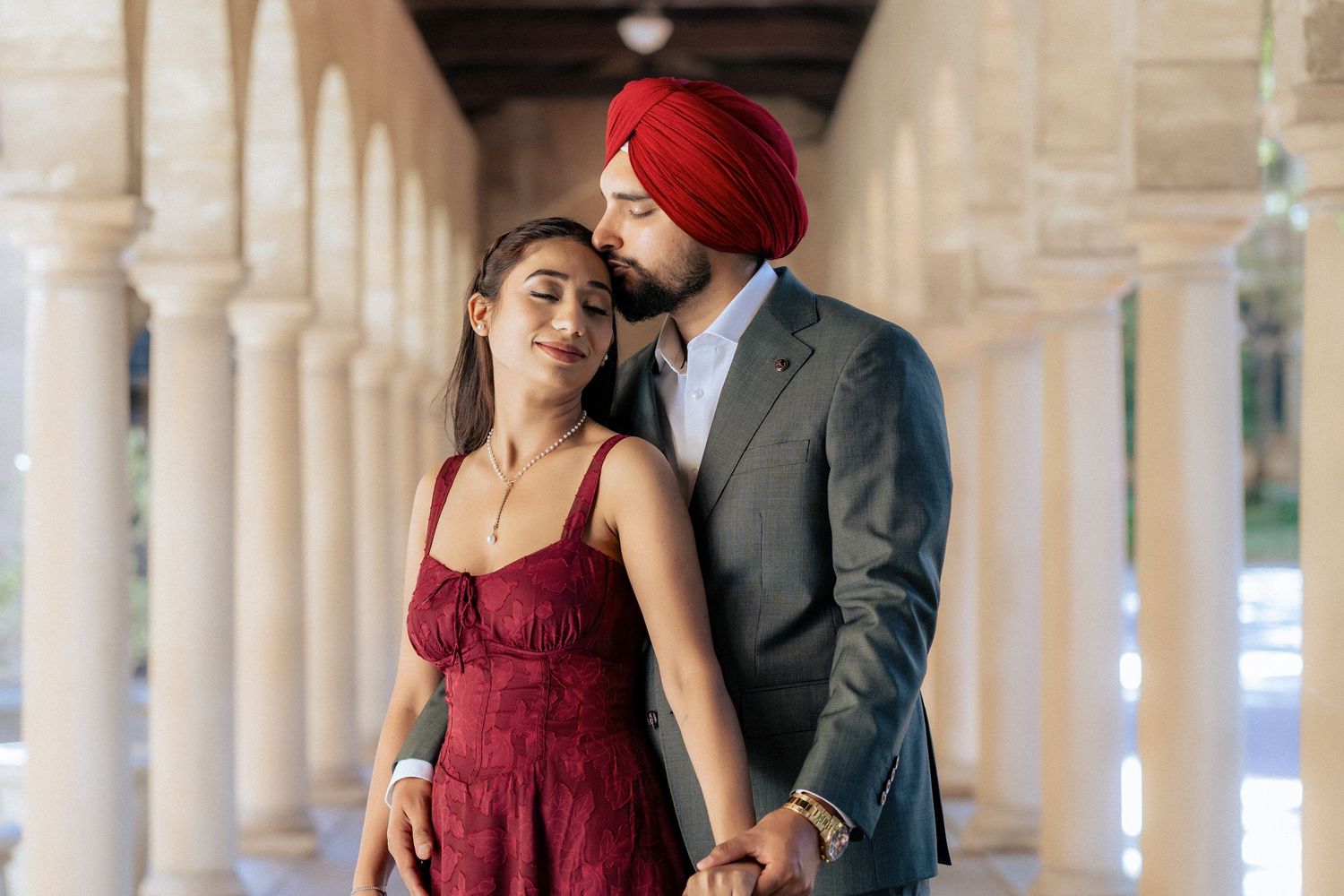 A Sikh couple in traditional attire shares a romantic moment under elegant arched columns.