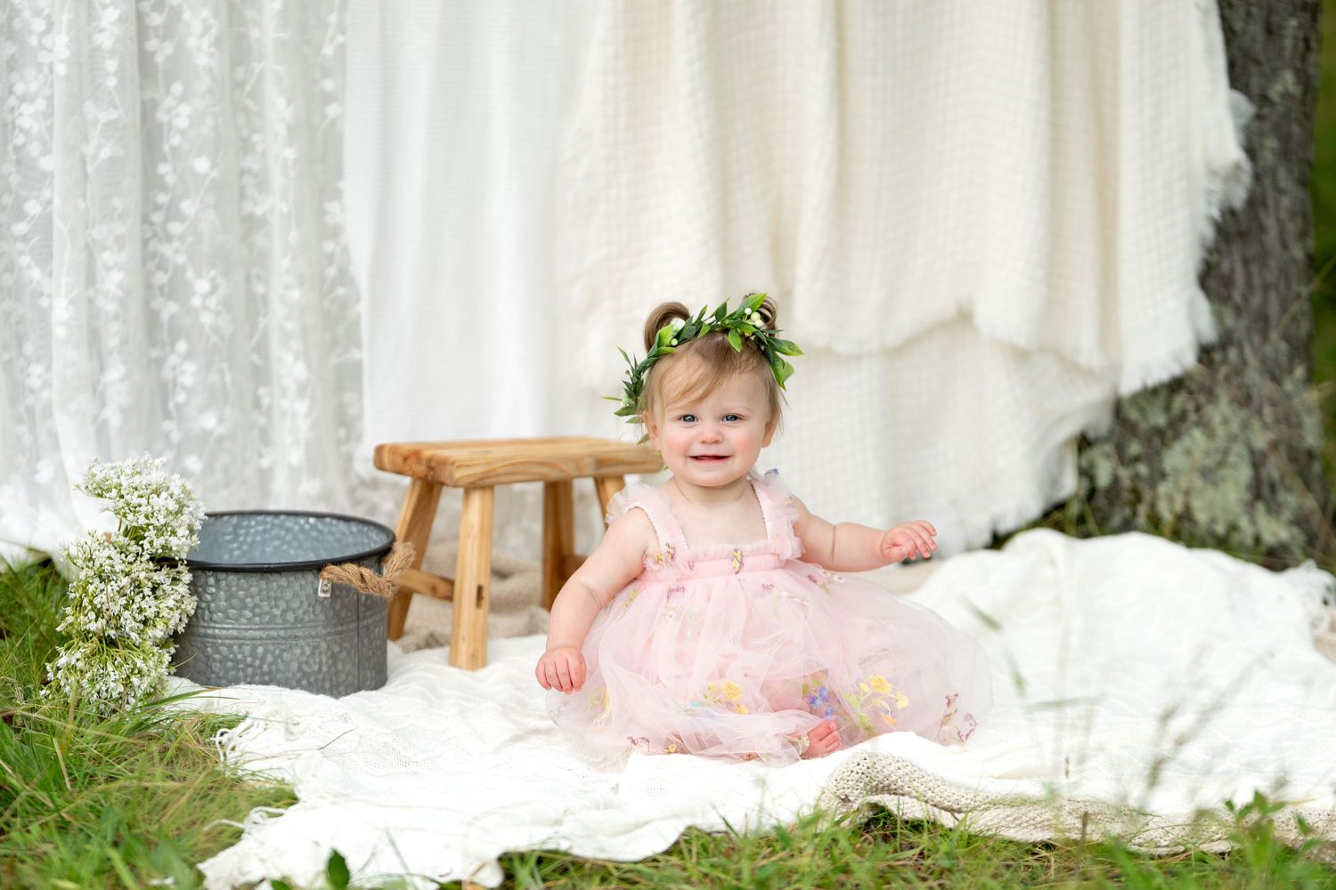 Smiling baby girl in pink dress and floral crown sits on white blanket outdoors in a charming photo shoot.