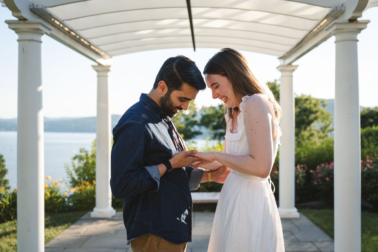 Joyful couple look at ring and embrace after proposal on cliff overlooking Hudson River and mountains during golden hour, under pergola.