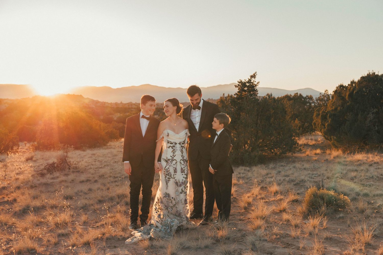 Wedding party of four standing in golden field with mountains and dramatic sunset in background.