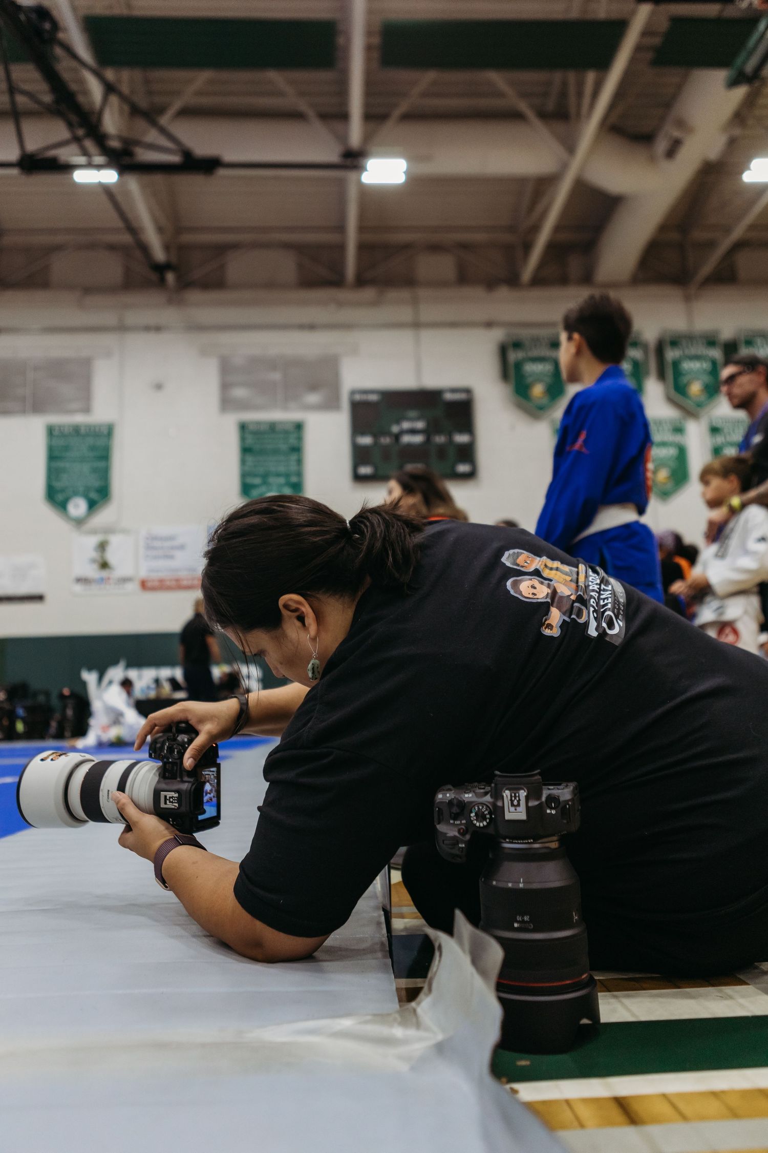 Photographer crouching at mat level with Canon cameras at an indoor jiu-jitsu tournament in a green gymnasium.
