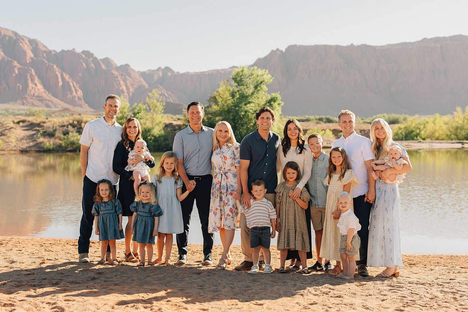Large family group portrait on sandy riverbank with red rock mountains and green trees in background.
