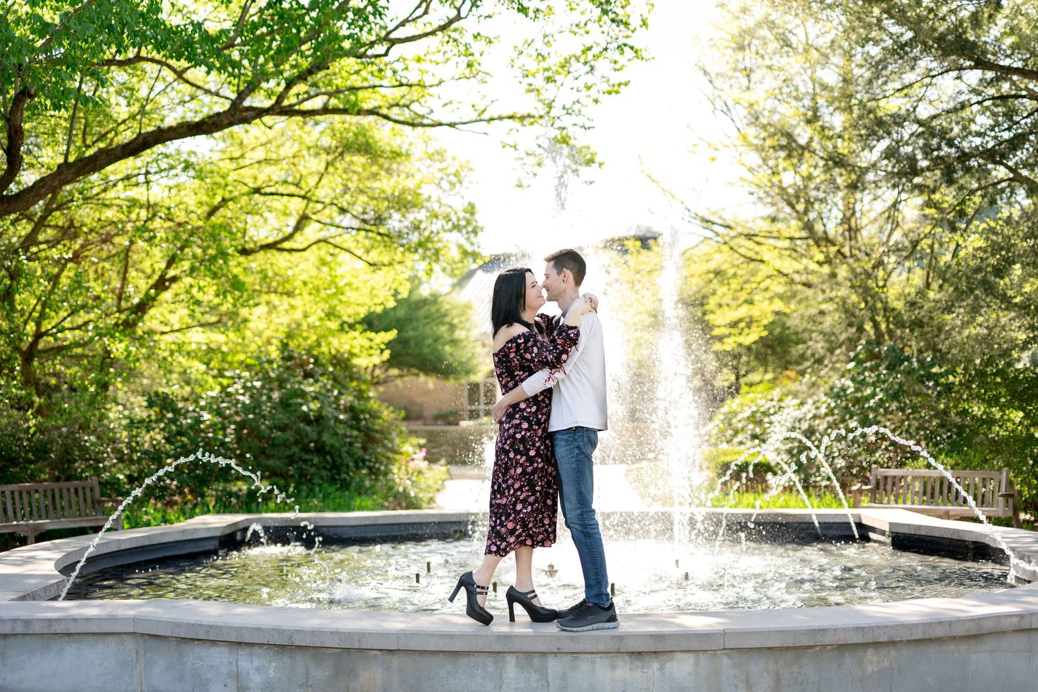 Couple kissing in front of a beautiful garden fountain surrounded by lush green trees in sunlight.