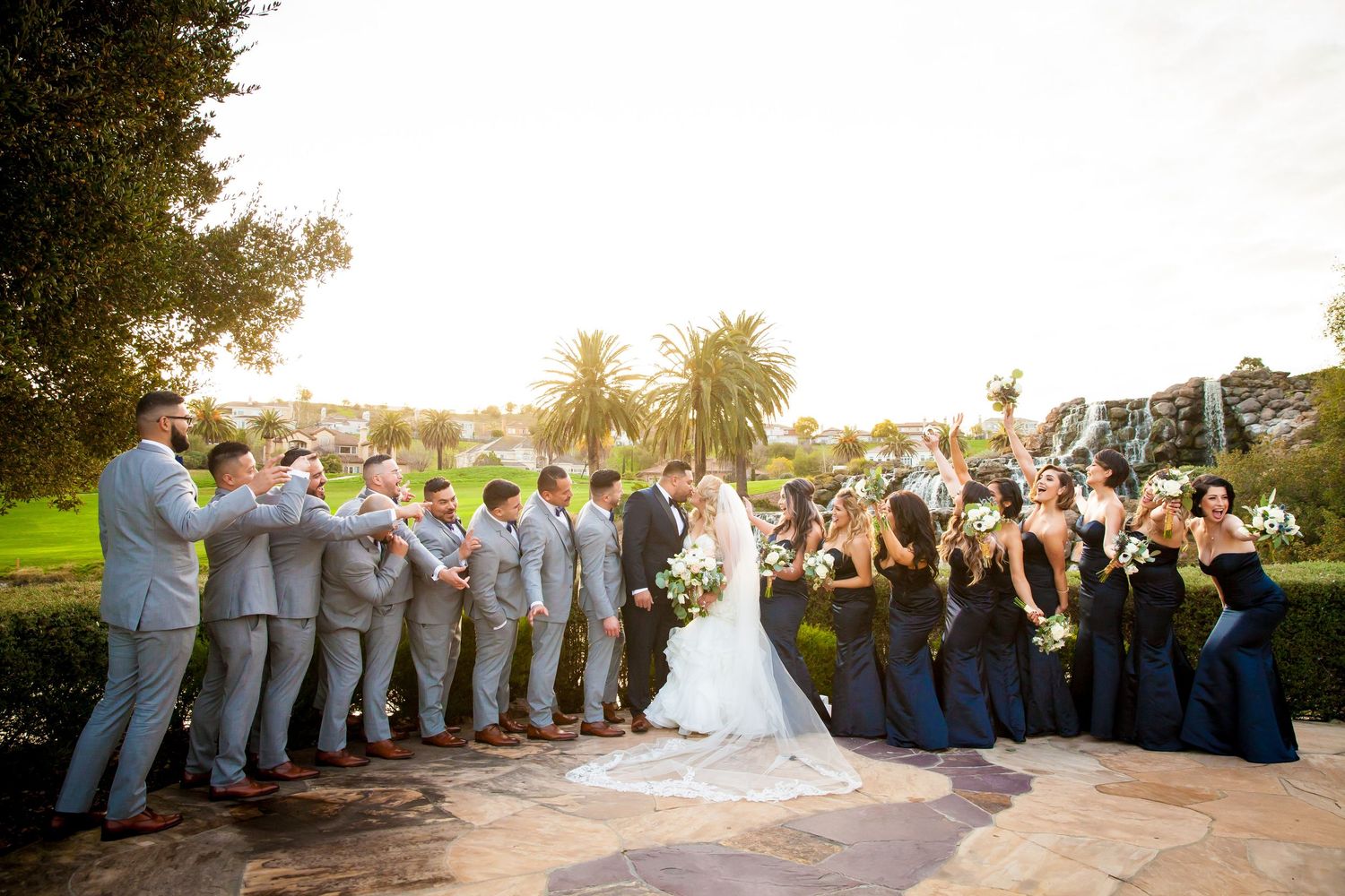 Wedding party members line up in a creative formation at sunset for a group photo at an outdoor venue.