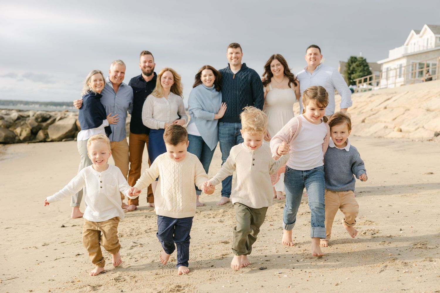 Large family posing on sandy beach with children holding hands in front, adults standing behind, coastal home in background.
