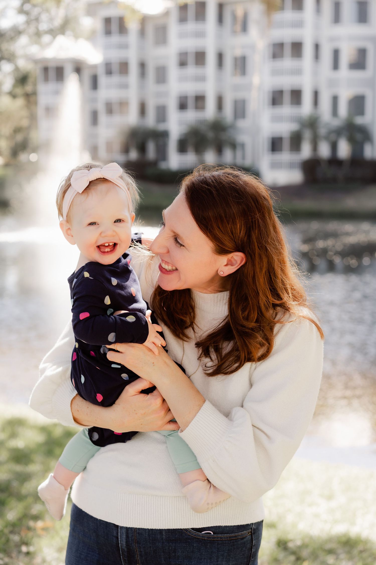 A parent in a cream sweater shares joyful moments with a baby wearing a navy outfit by a waterfront building.