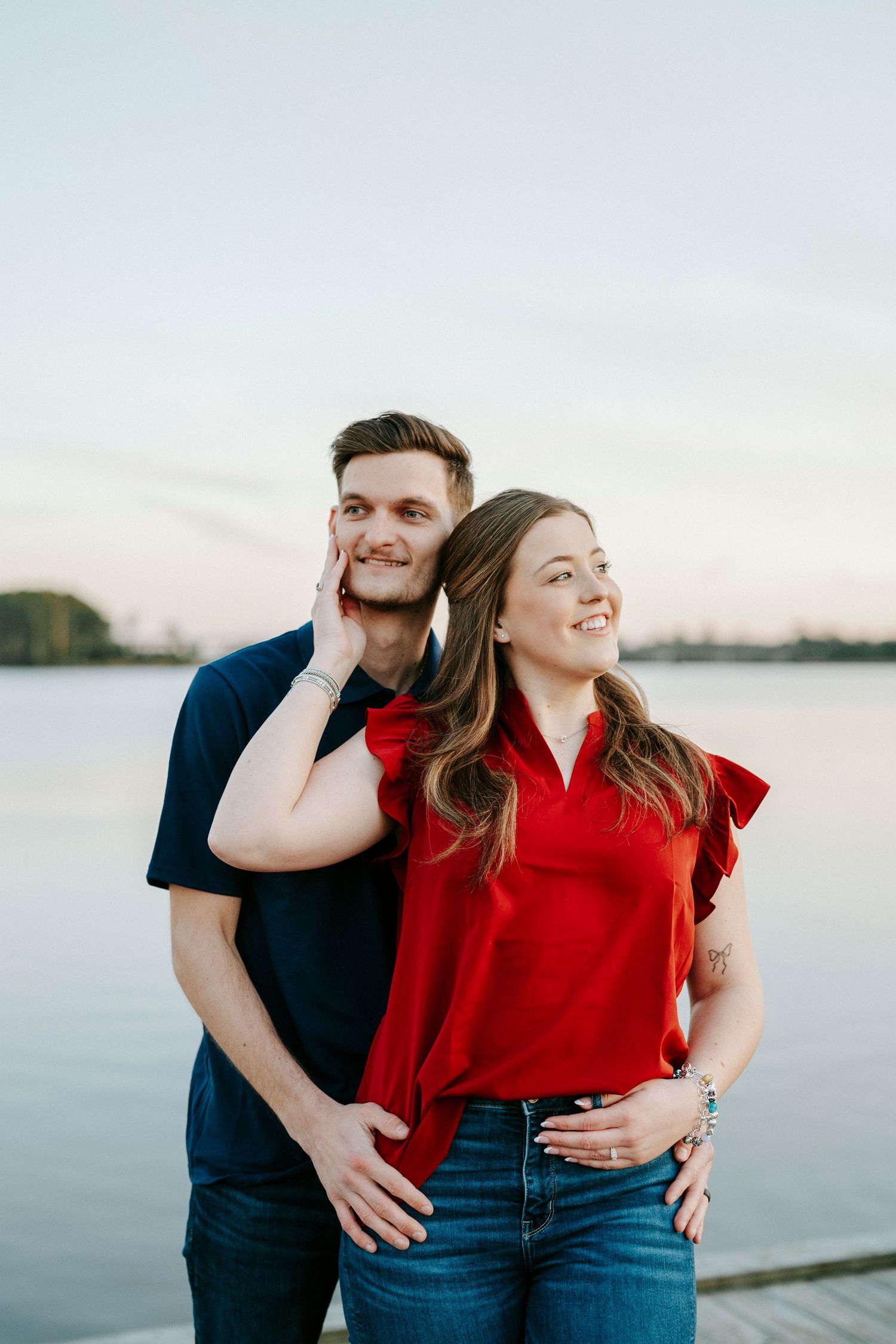 A couple in navy and red outfits share intimate moments by a waterfront during a sunset photo session.