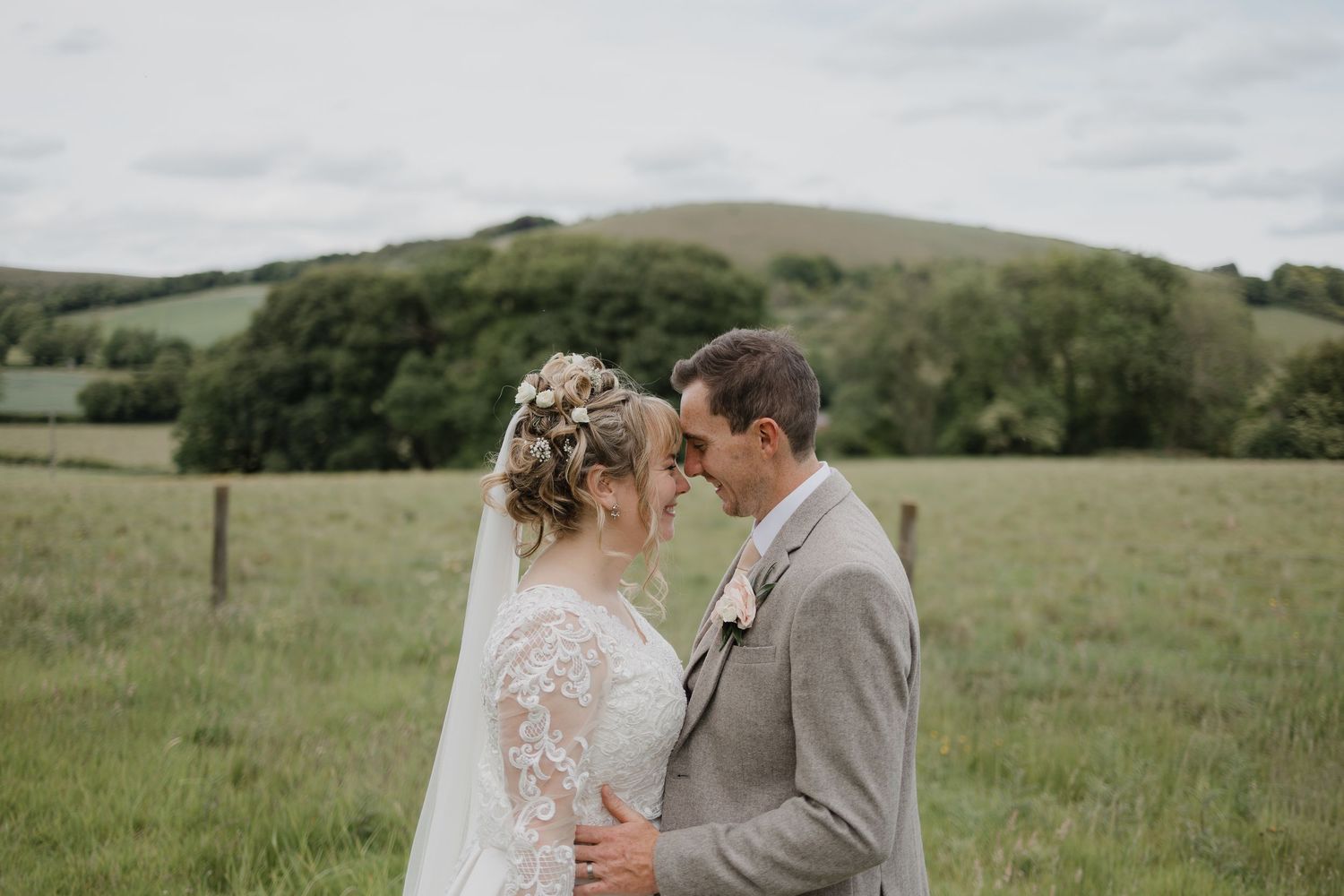 A couple shares a romantic moment in a green rolling meadow during their rustic outdoor wedding celebration.