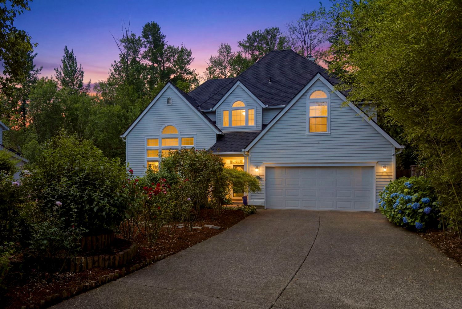Two-story light blue home with attached garage photographed at dusk with warm interior lighting and lush green trees.