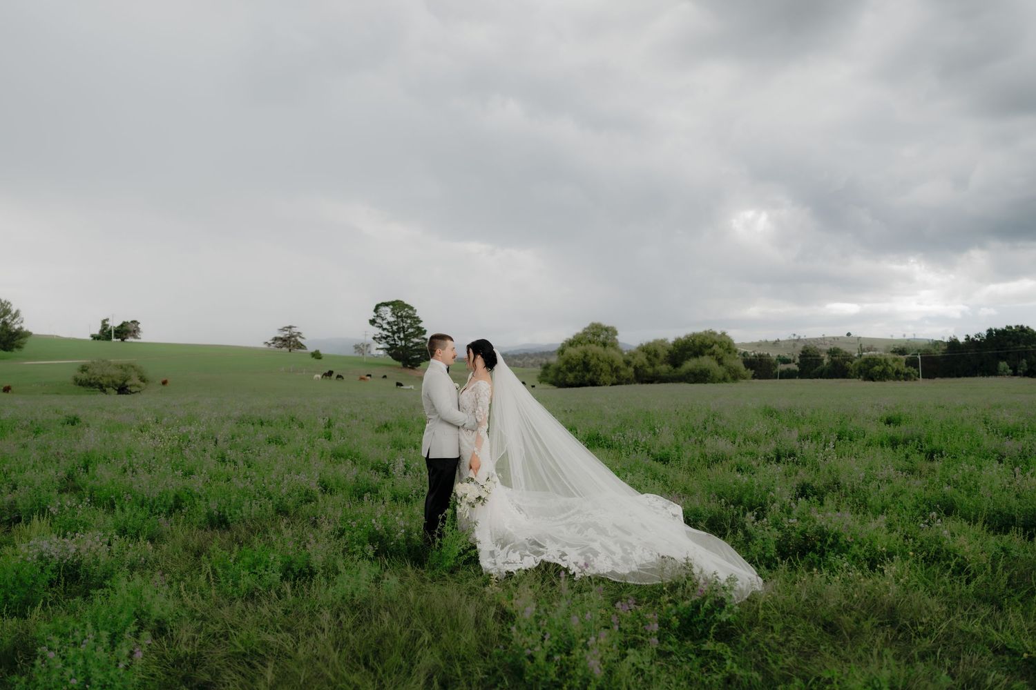 A bride and groom share a romantic kiss in a lush green field under dramatic cloudy skies, her long veil trailing behind.