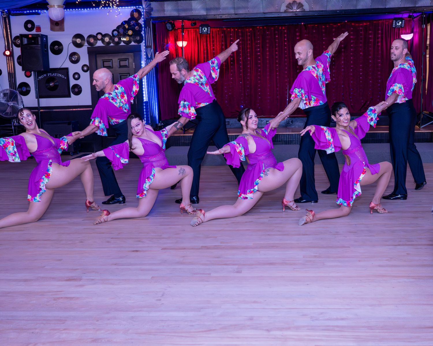Latin dance performers in purple costumes strike synchronized poses on a ballroom dance floor at showcase event
