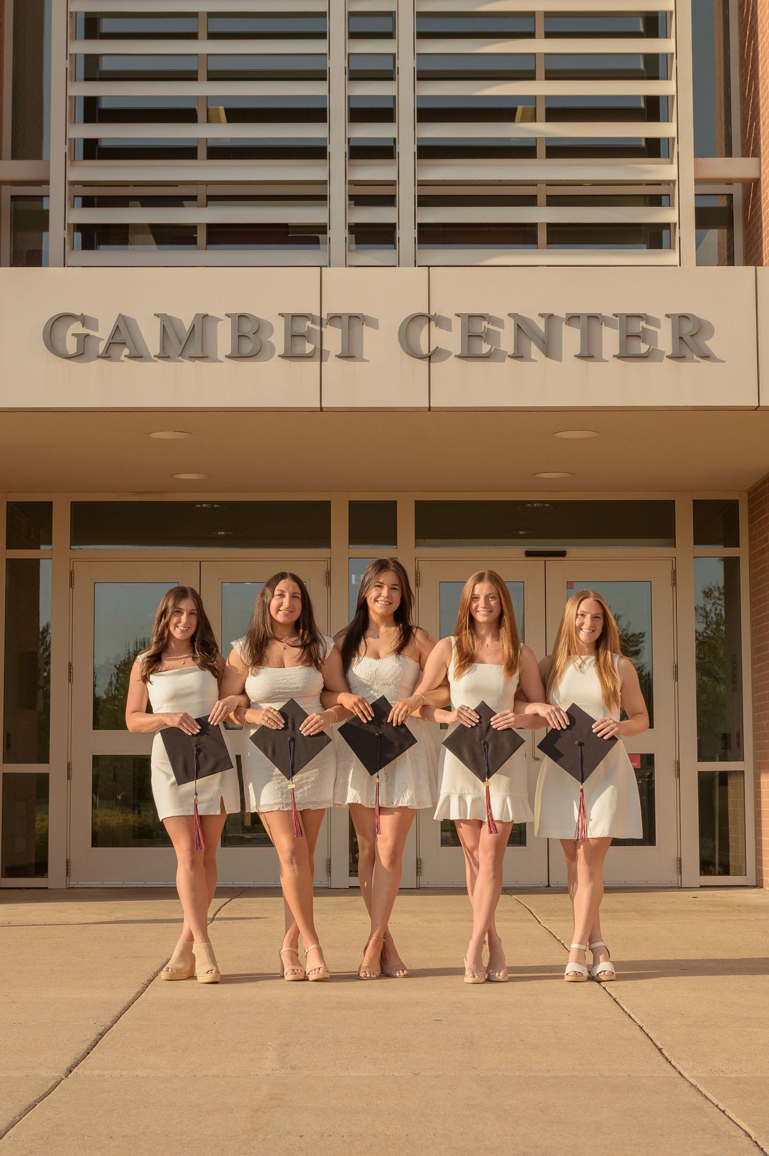 Five female graduates in white dresses hold caps outside the Gambet Center building.