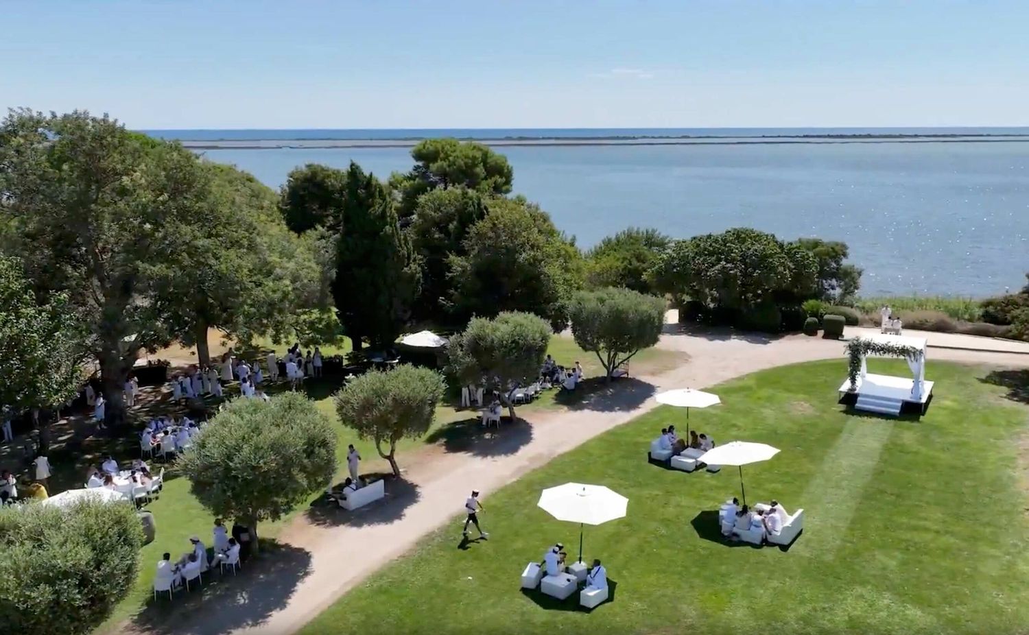 Aerial view of an elegant outdoor waterfront event with guests on lush green lawn, white umbrellas, and ocean backdrop.