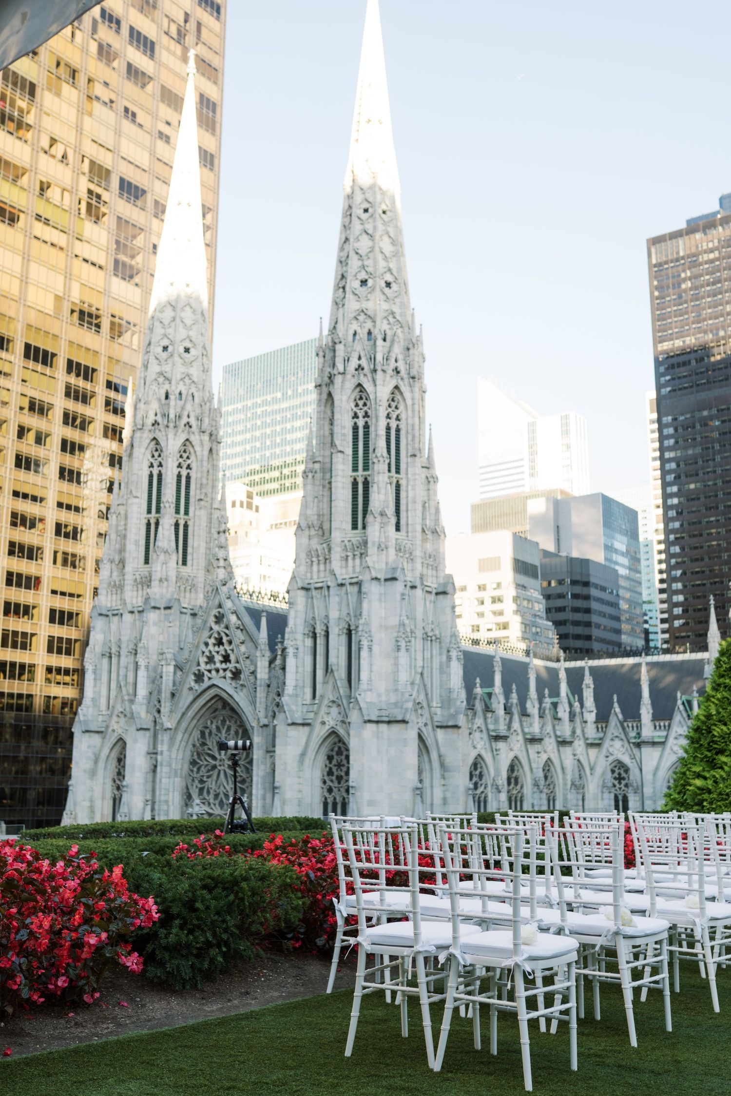 Outdoor wedding ceremony setup with white chairs on green lawn, St. Patrick's Cathedral and NYC skyscrapers in background.