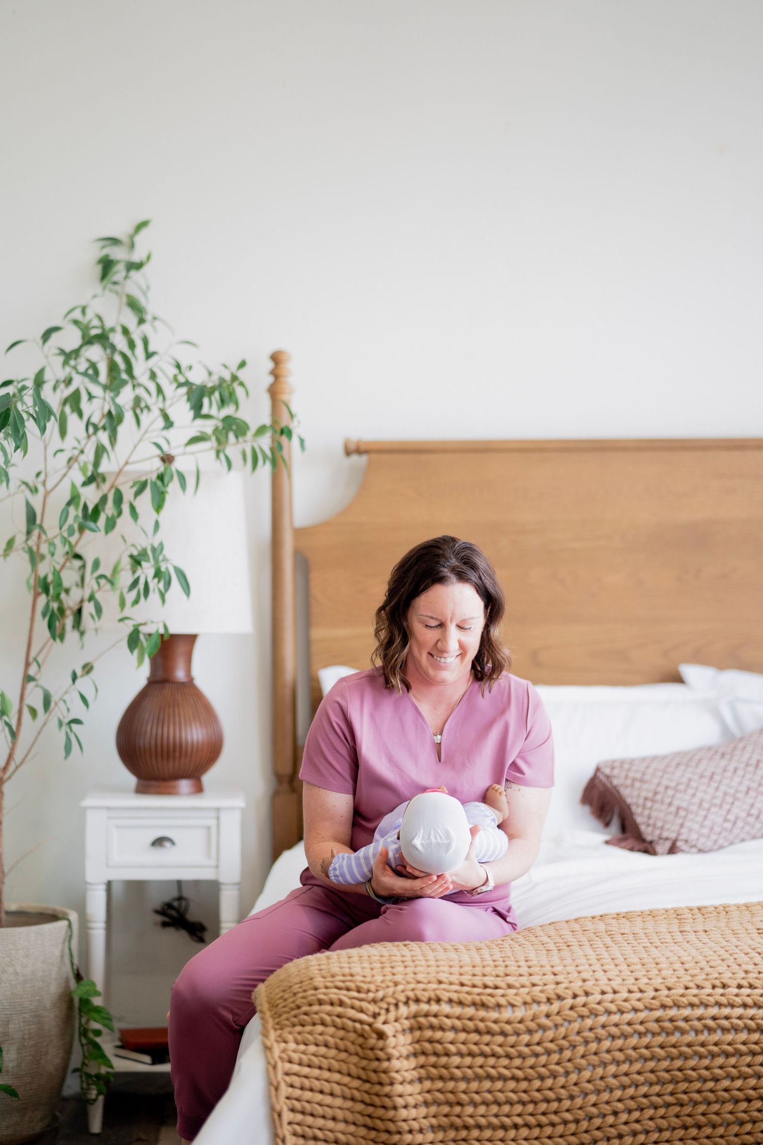 Nurse in pink scrubs sitting on bed holding newborn baby, looking down lovingly in bright bedroom.
