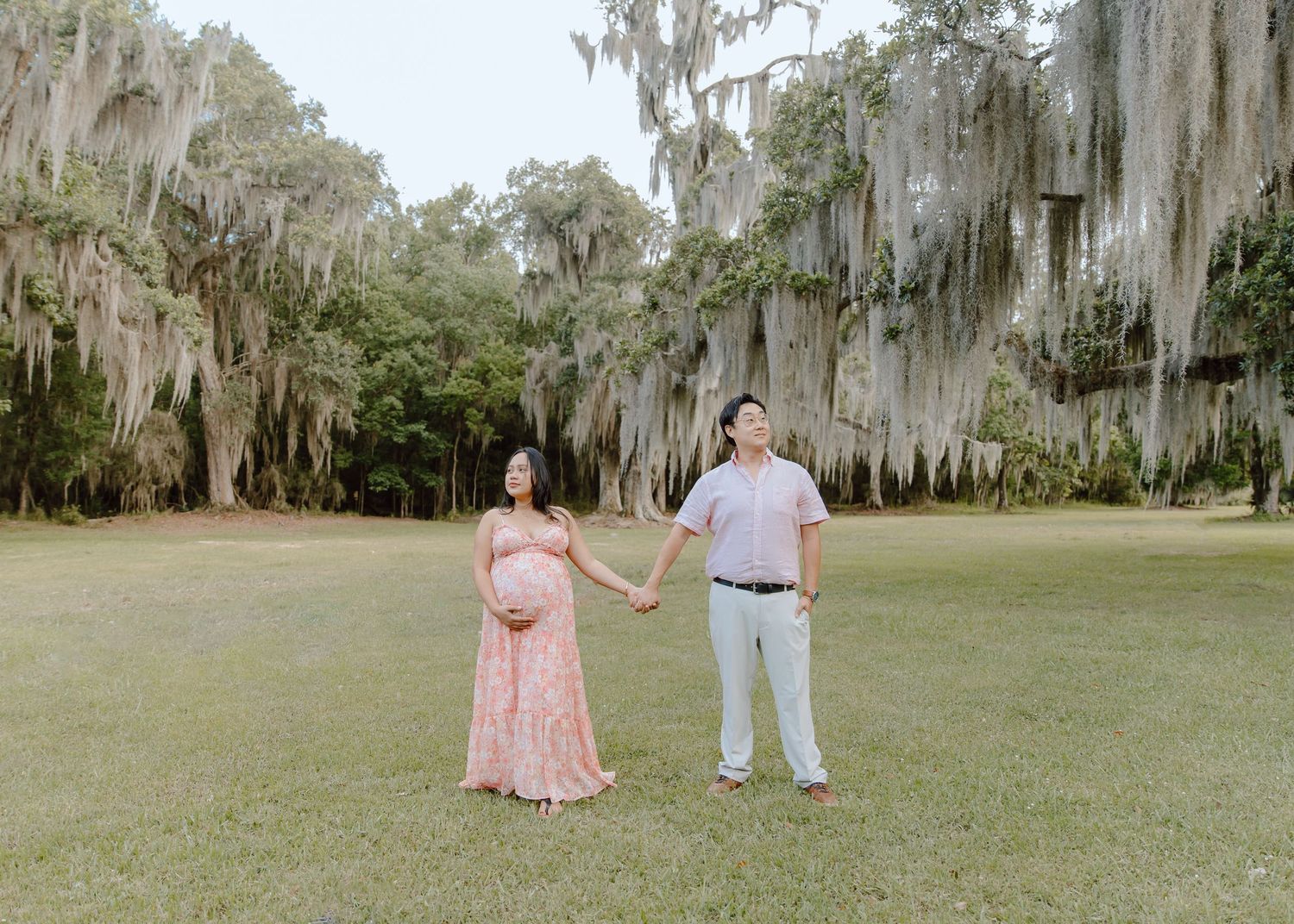 Pregnant couple holding hands in a lush green park with Spanish moss-draped oak trees during maternity photoshoot.
