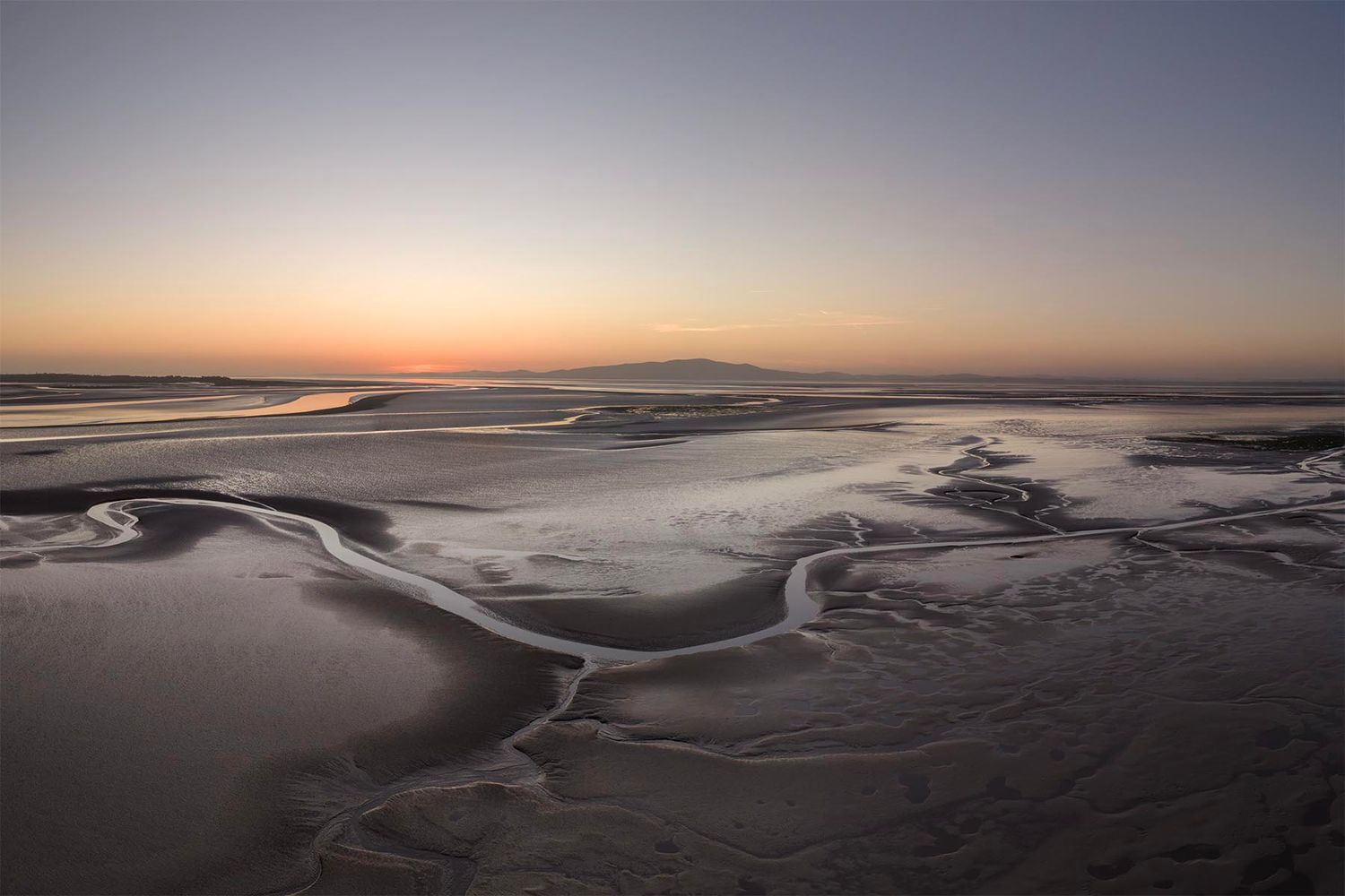Aerial view of tidal flats at sunset with winding water channels across sandy beach and distant mountain silhouette.