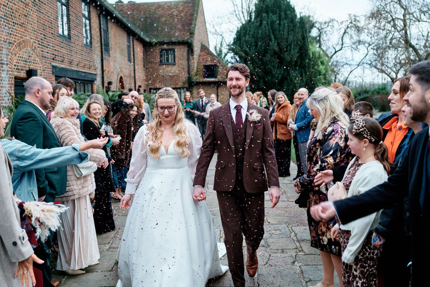 Bride and groom walk hand in hand through a confetti shower, guests lining the path at the Tudor Barn in Eltham.