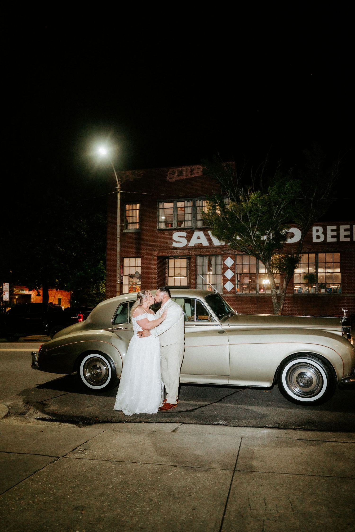 Newlyweds kiss beside a vintage silver car at night outside Salvage Beer venue.