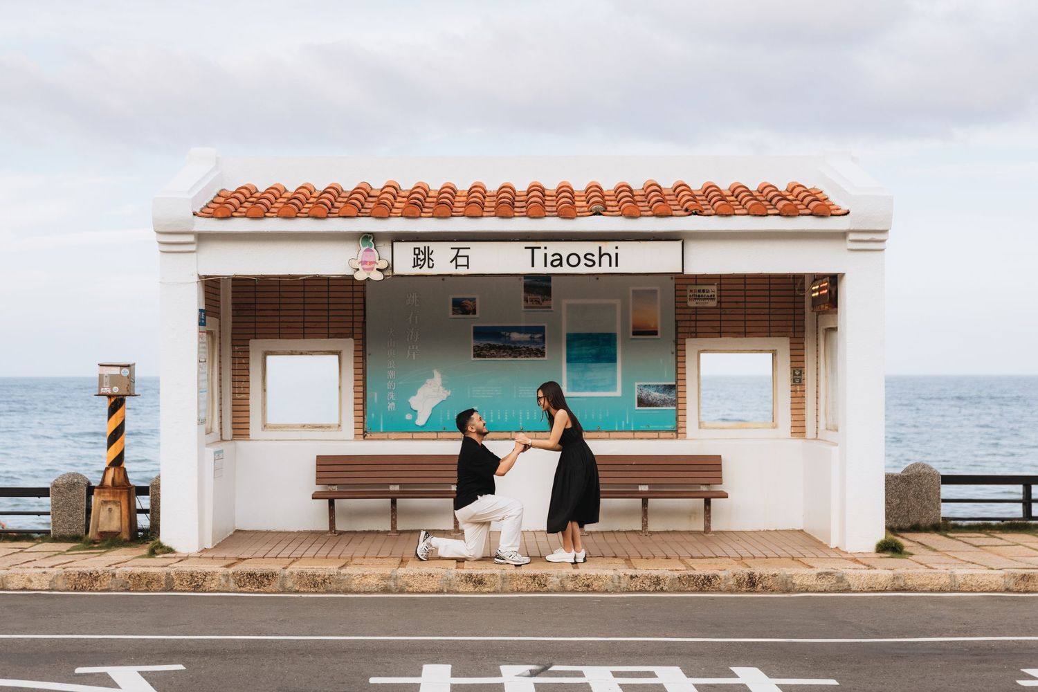 Couple at Tiaoshi train station in Taiwan, man proposing on one knee with ocean backdrop.