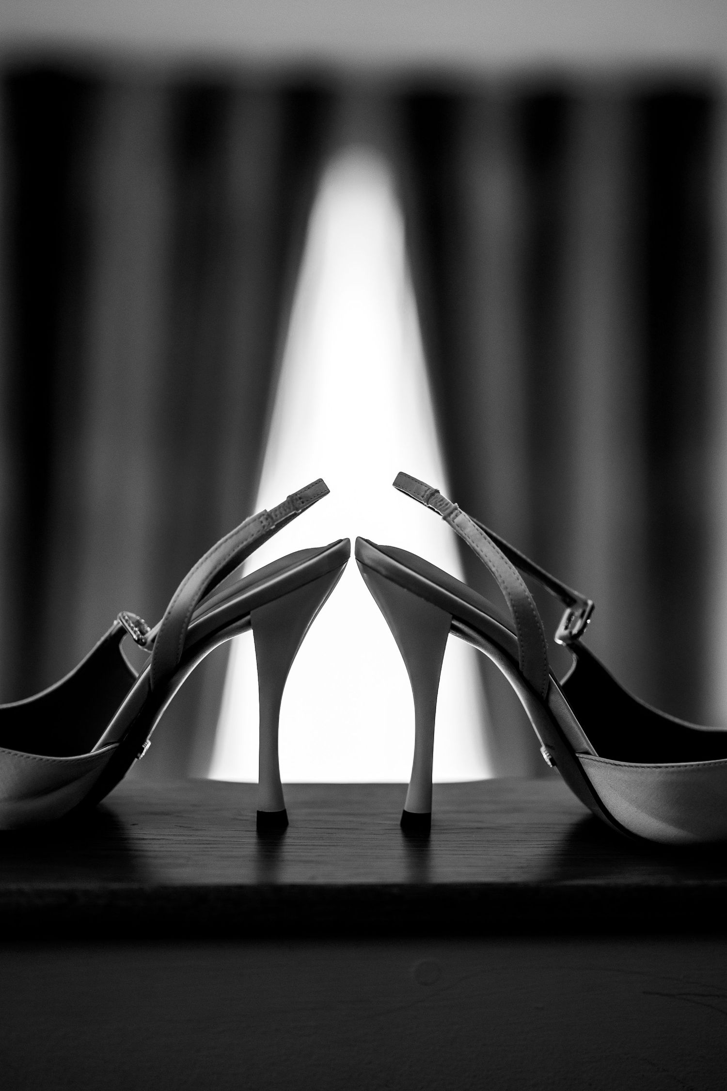 Black and white photo of elegant high heel slingback shoes on a dark surface with dramatic curtain backdrop.