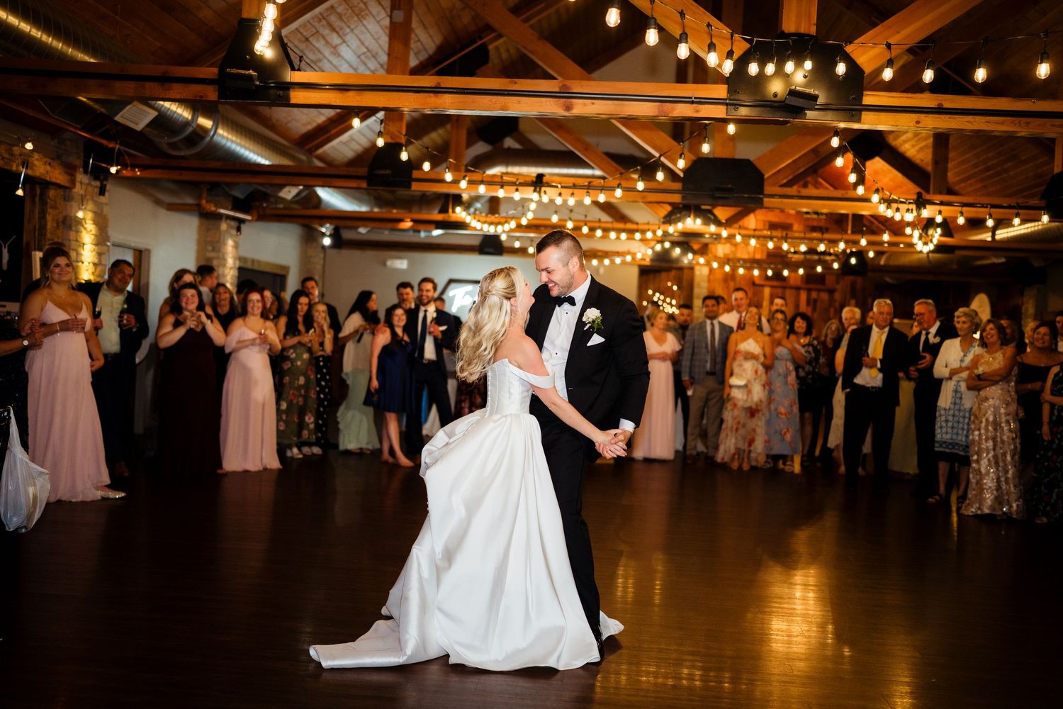 Bride in white off-shoulder gown smiles joyfully as groom kisses her cheek during romantic outdoor wedding portrait.