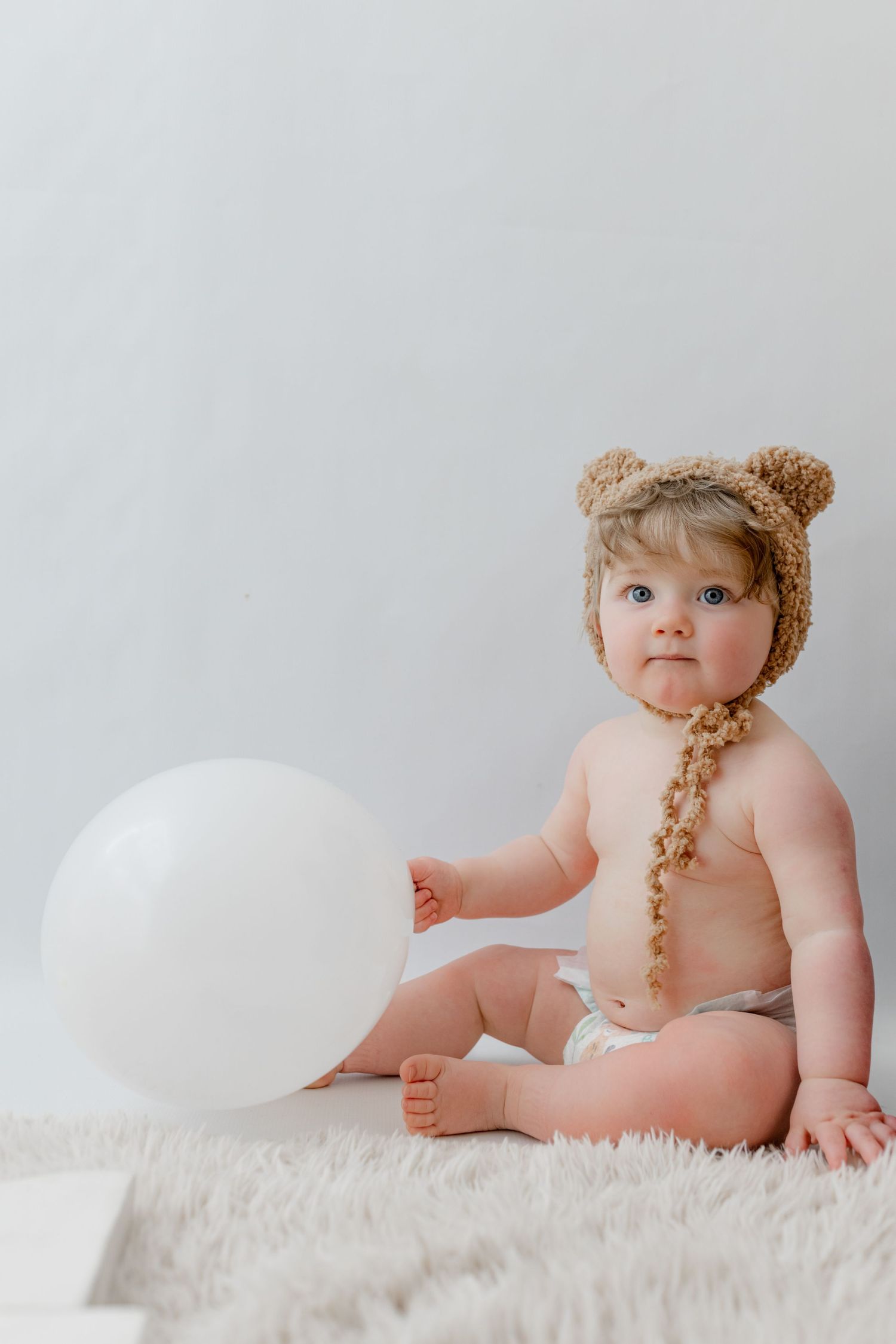Baby wearing brown bear hat sits on white fluffy rug with balloon in minimalist portrait setting.