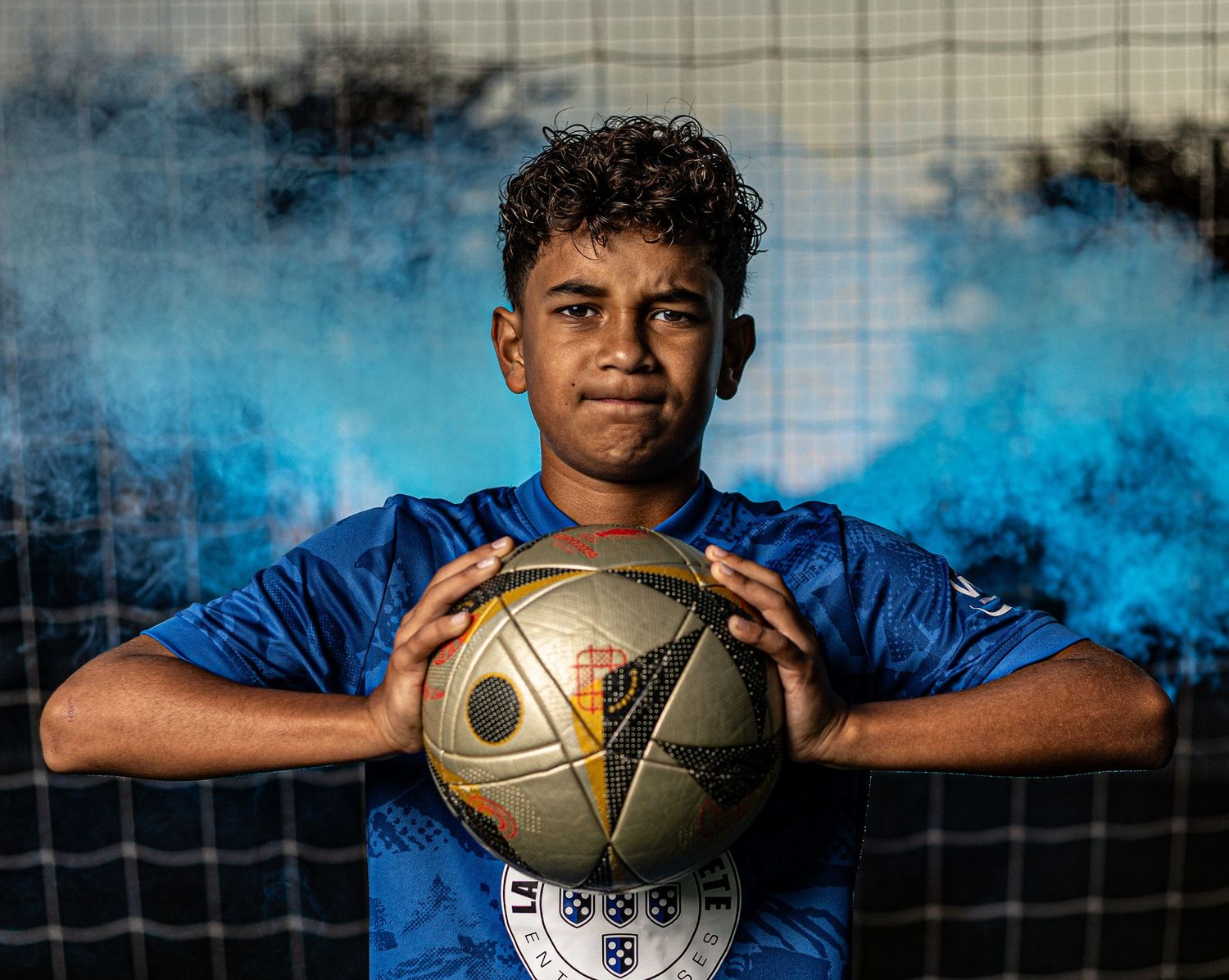Young soccer player in blue jersey holds a football in front of a net with blue smoke in the background.