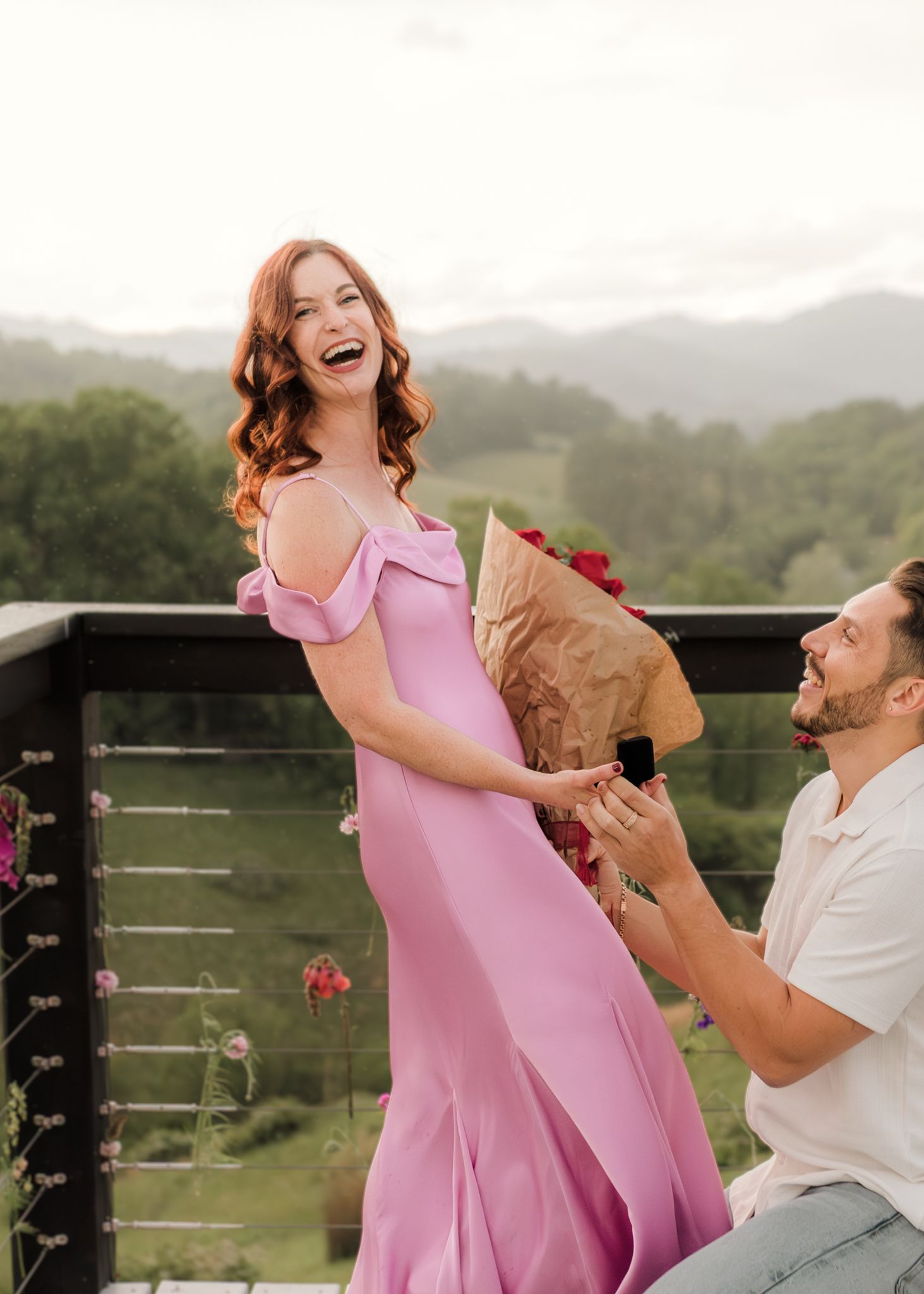 Man proposes to laughing woman in pink gown on scenic mountain balcony, holding flowers and ring box.