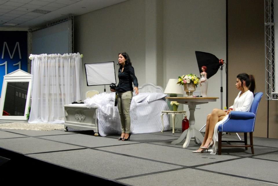 Two women on a stage with wedding decor, white drapes, floral arrangements, and photography equipment at a bridal expo.