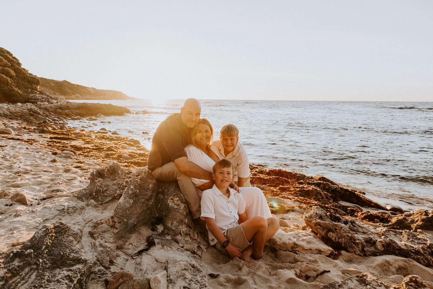 Family of four posing on rocky beach at golden sunset, smiling together by the ocean shore.
