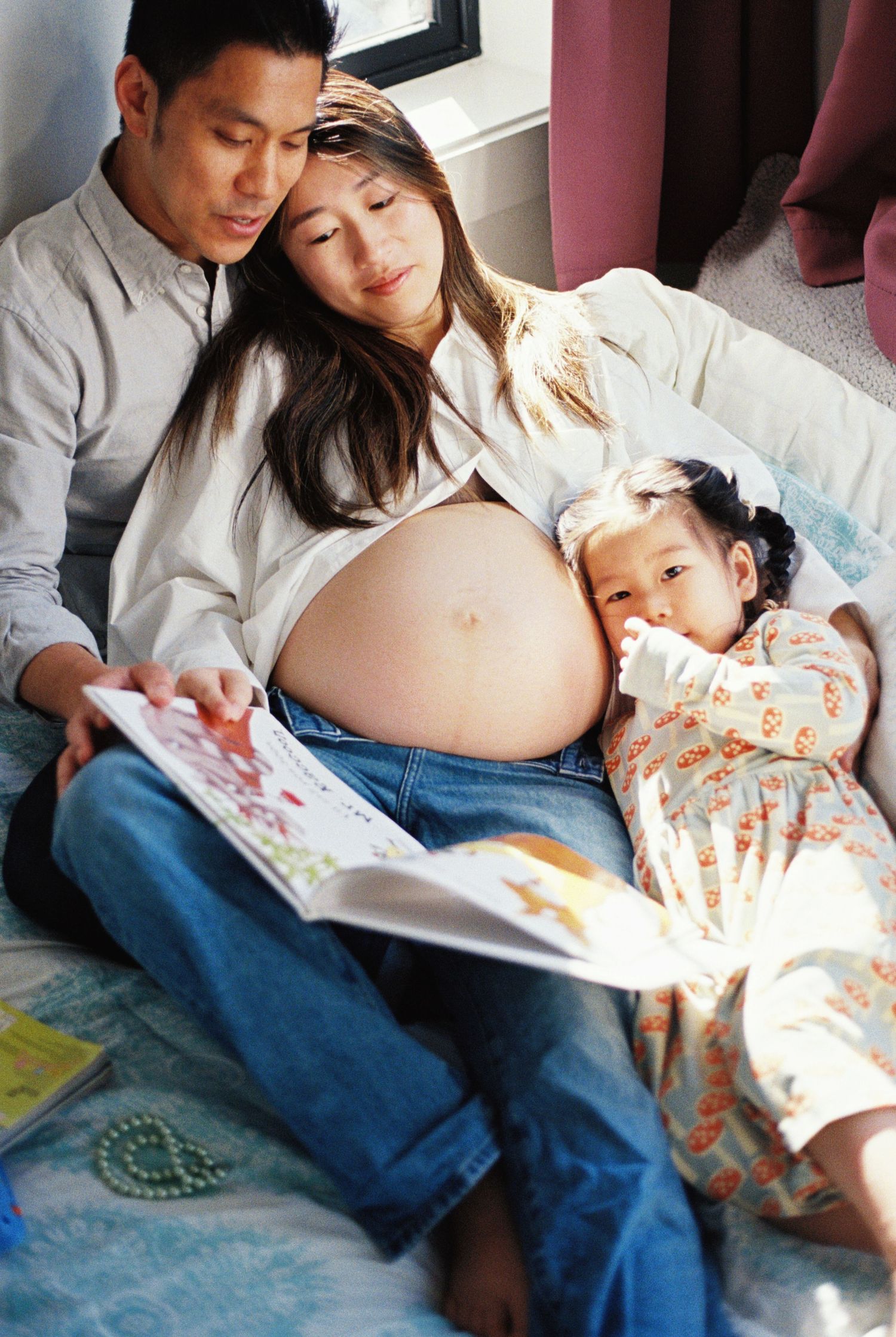 Pregnant Asian woman reading a book with her husband and young daughter, enjoying a cozy family moment together.