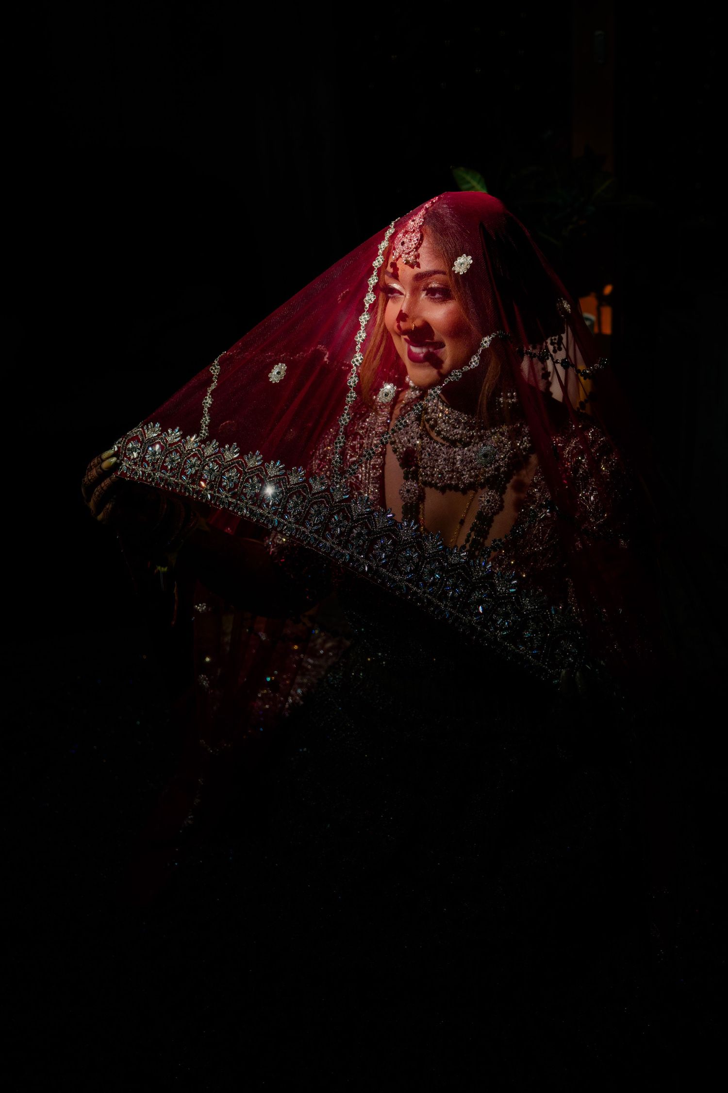 Indian bride in red dupatta with silver jewelry, dramatically lit against dark background.