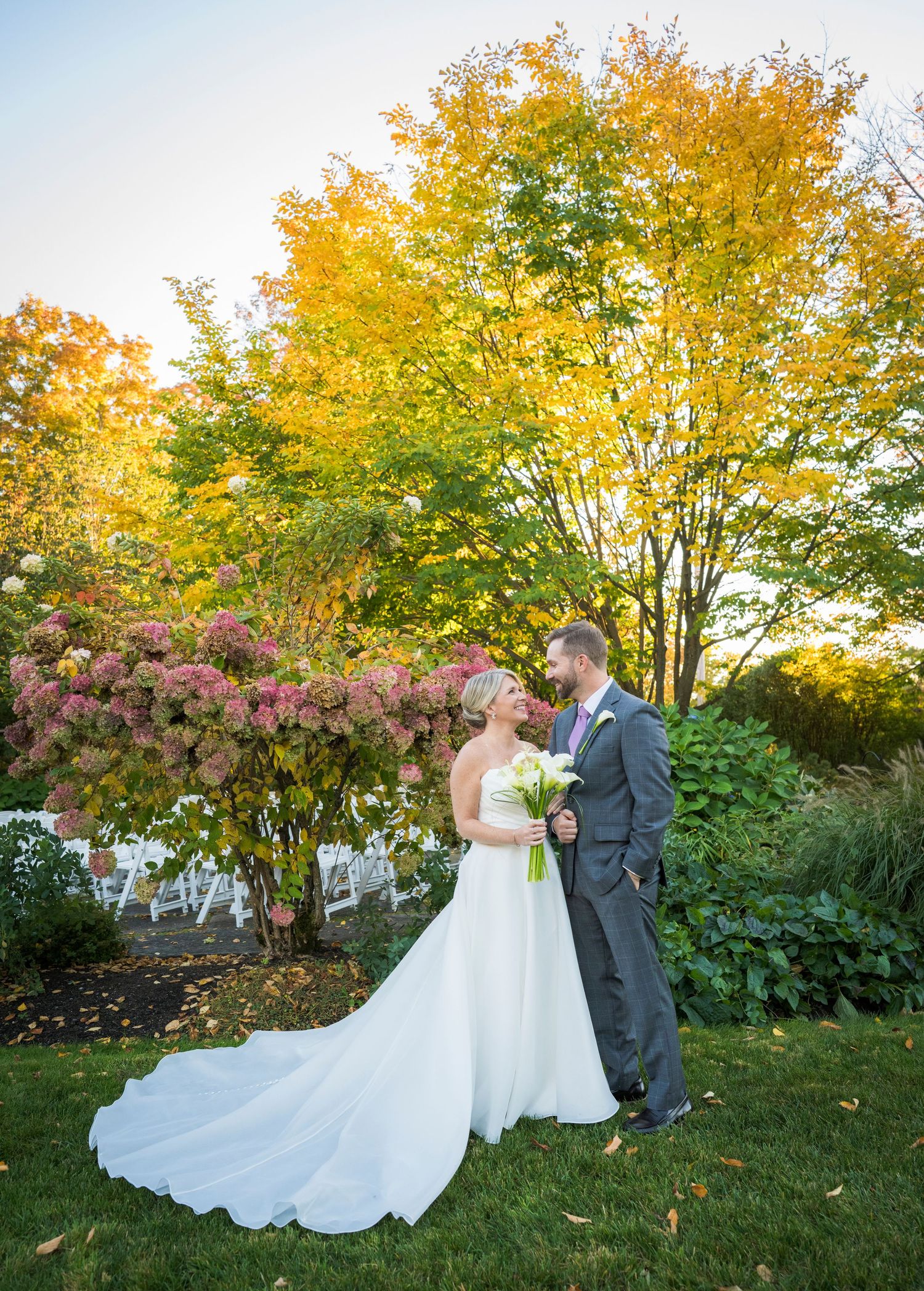Bride and groom share a romantic sunset kiss outdoors, surrounded by lush greenery and tall ornamental grasses.