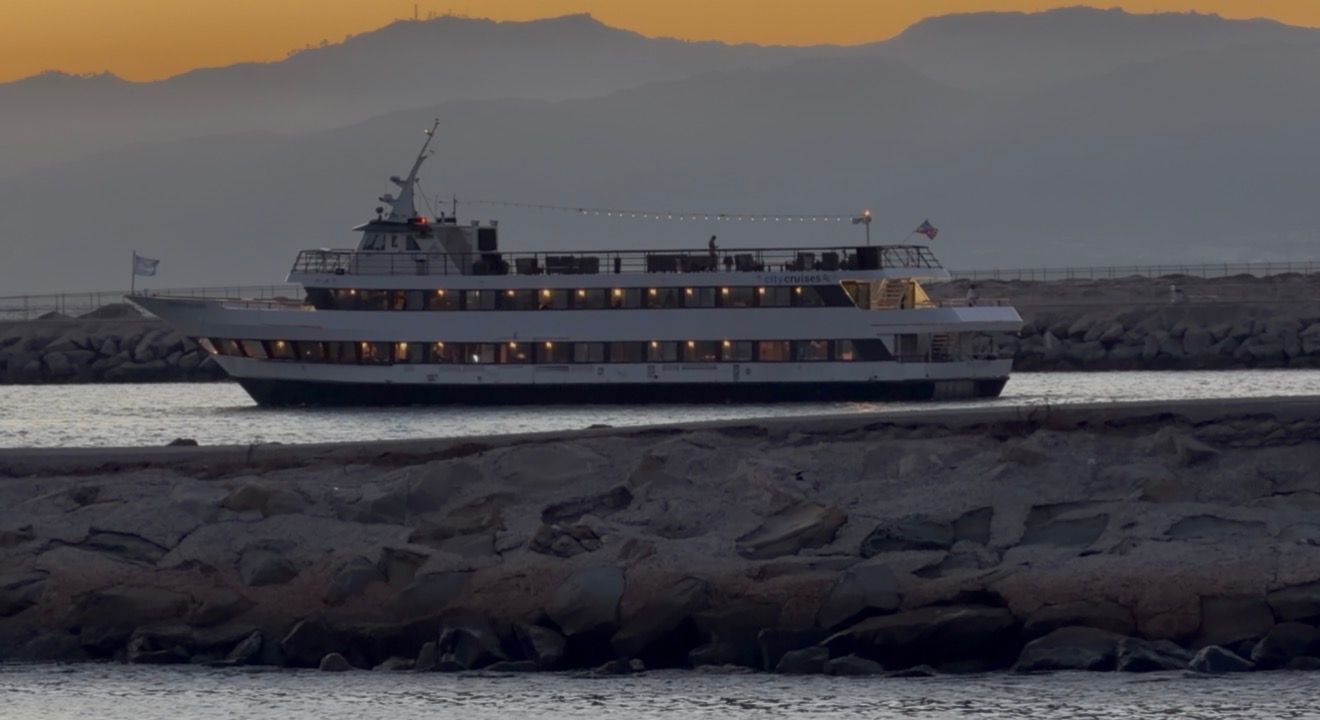 A large cruise ferry boat sailing along a rocky shoreline at dusk with mountains in the background.