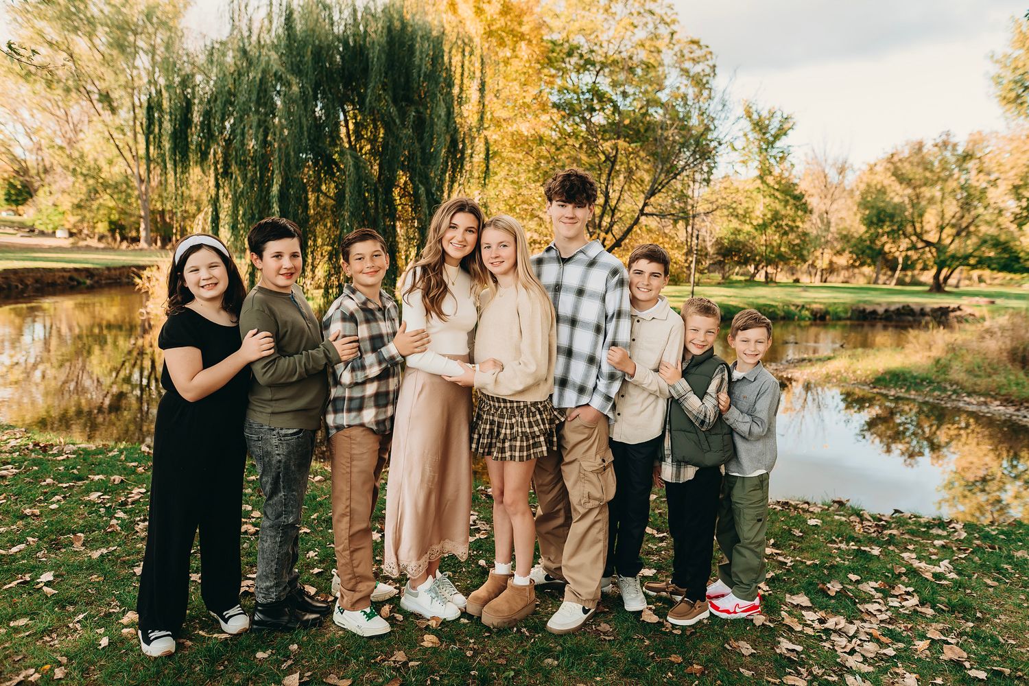 Group of eight children smiling together outdoors near a pond surrounded by autumn foliage.