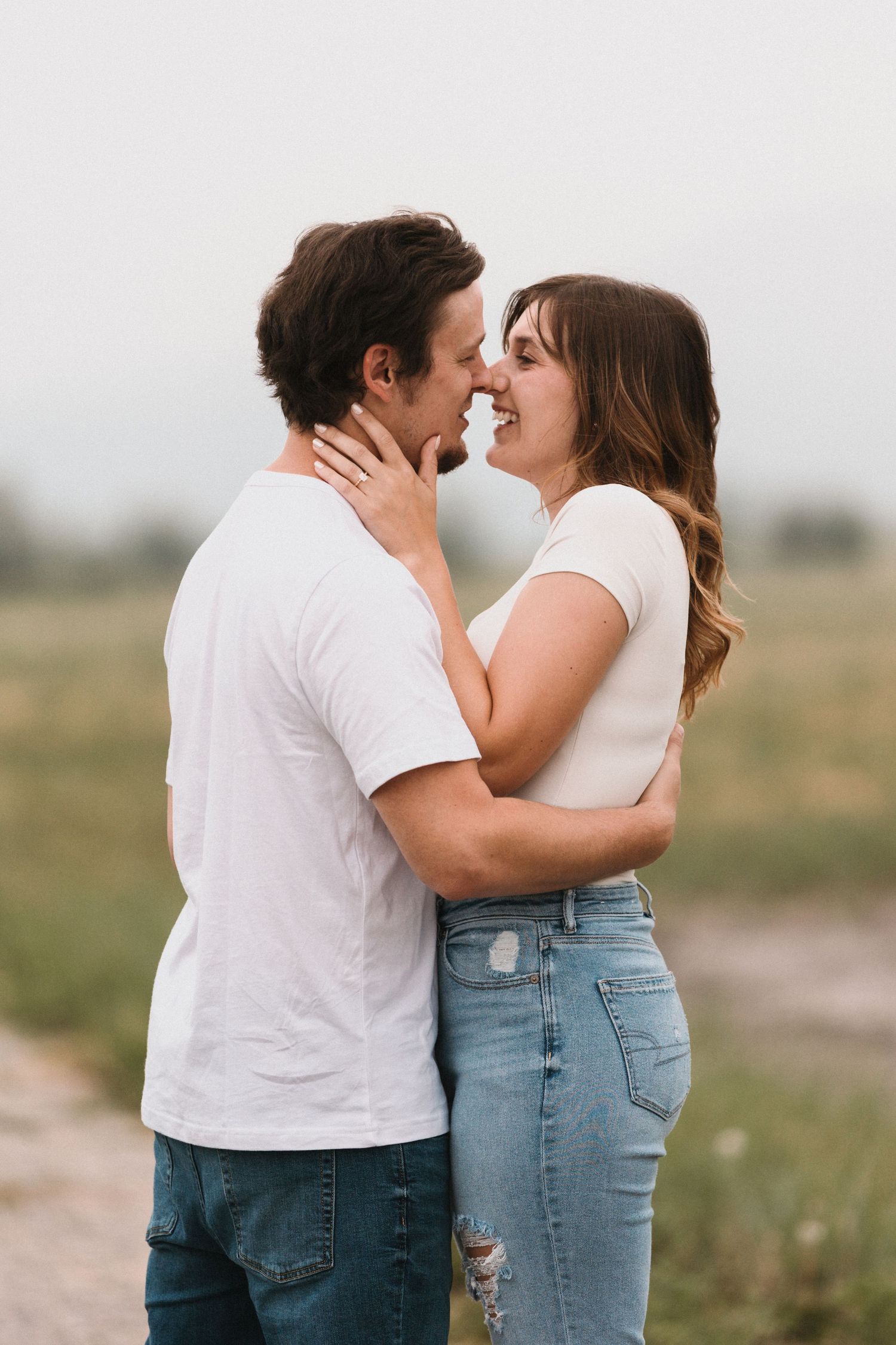 A couple in white tees and jeans share a romantic moment outdoors, smiling and about to kiss in a field.