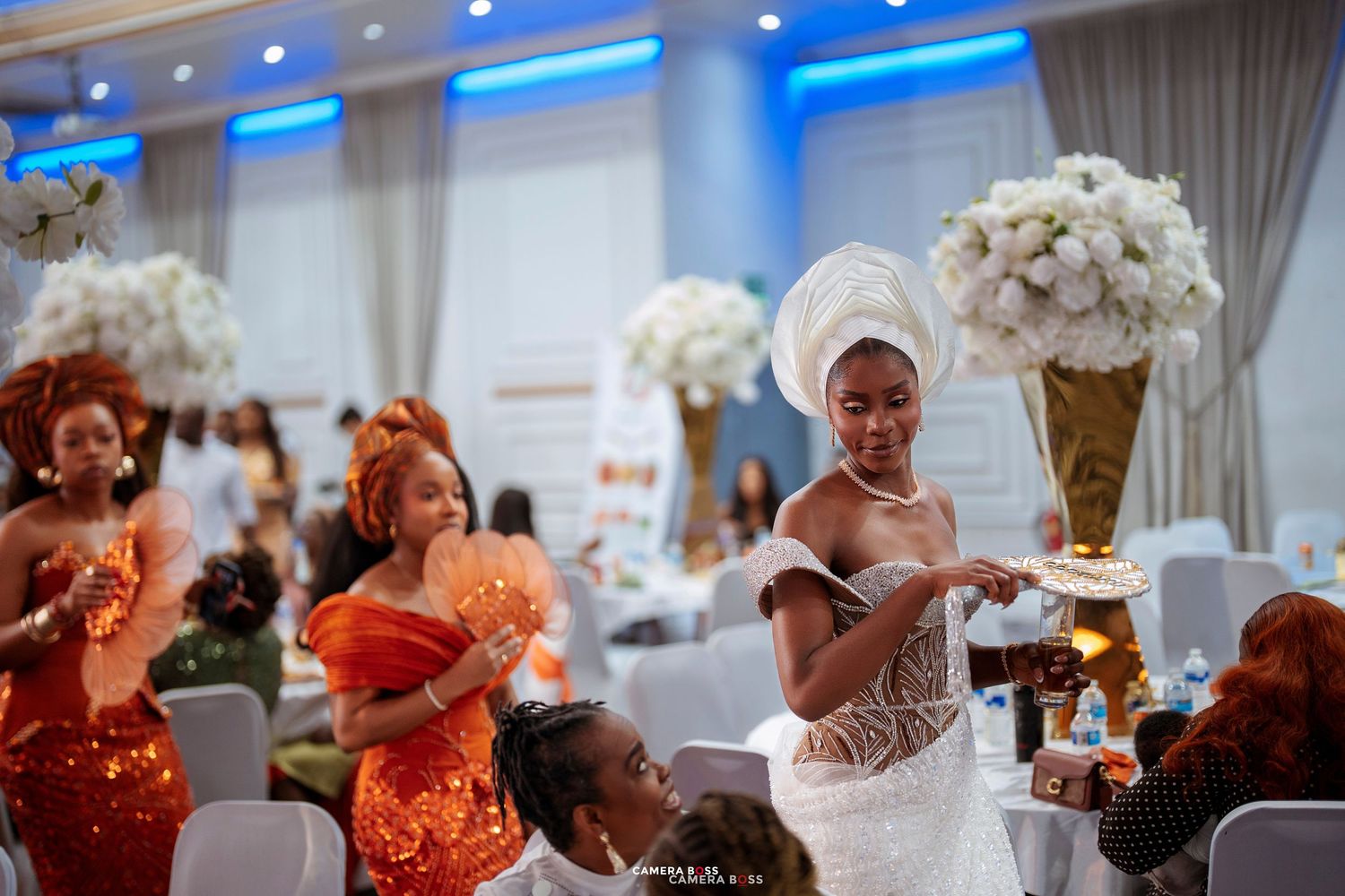 Elegant African bride in white lace gown and gele headwrap at a vibrant wedding reception with guests in colorful attire.
