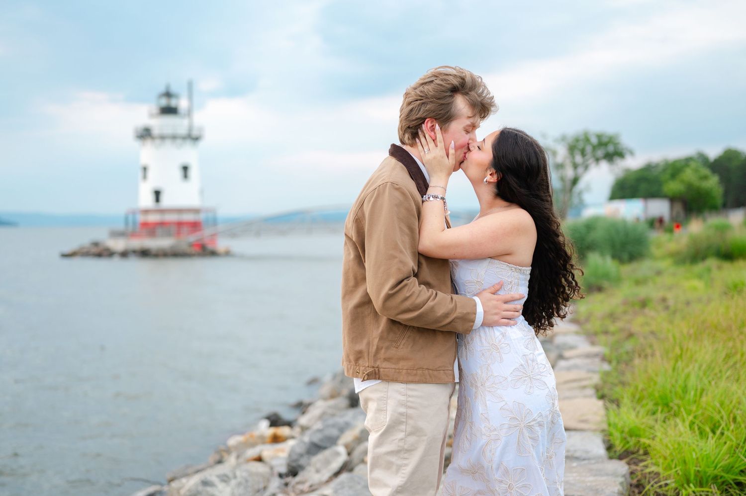 Couple near a lighthouse by the Hudson River, man kneeling on rocks proposing to woman in white dress.