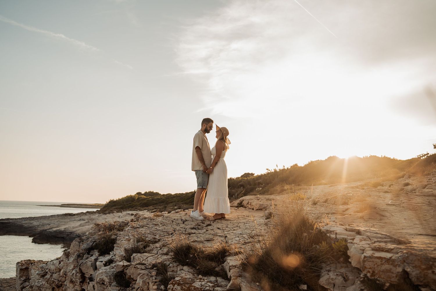 Couple sharing a romantic sunset kiss on rocky coastal cliffs with golden light and ocean views.