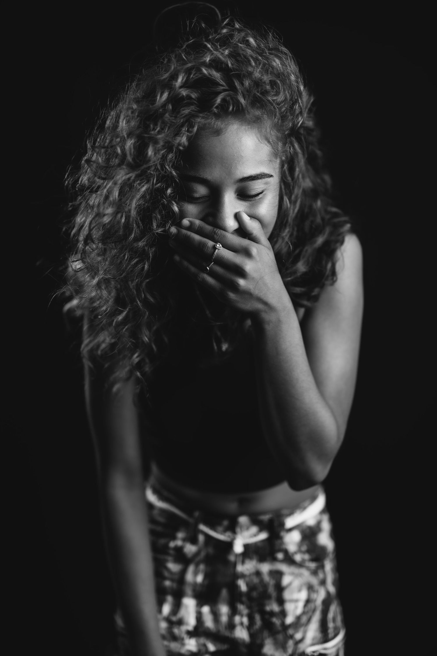 Artistic black and white portrait of someone with curly hair laughing behind their hands.