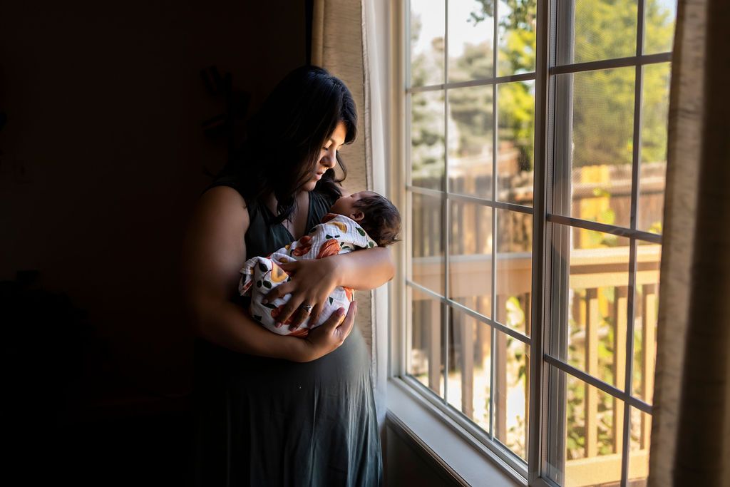 Woman stood next to a window, dark shadows behind her, she is cradling and looking down at a newborn baby