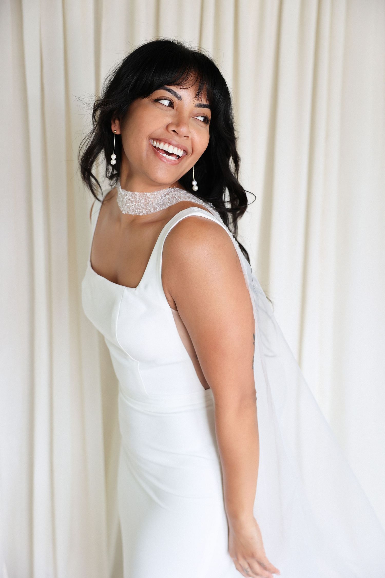 A radiant woman in a white wedding dress and sparkling choker smiles warmly against cream curtains.