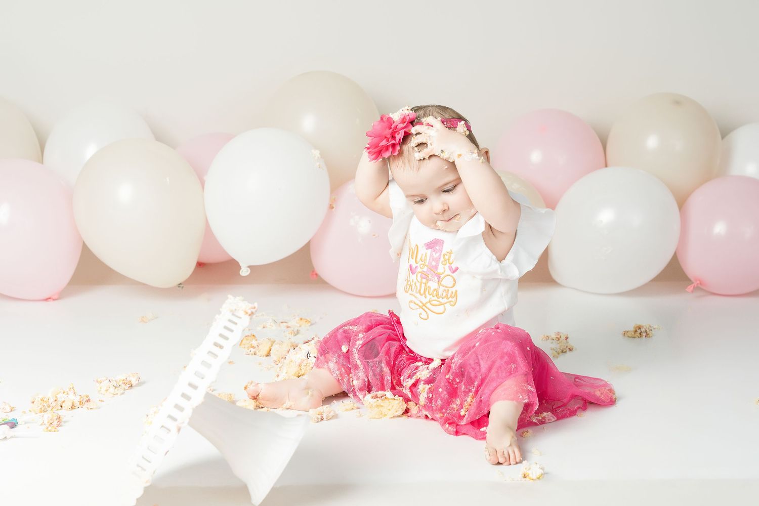 Baby sitting on floor with pink and white balloons for cake smash photo session wearing tutu and headband.
